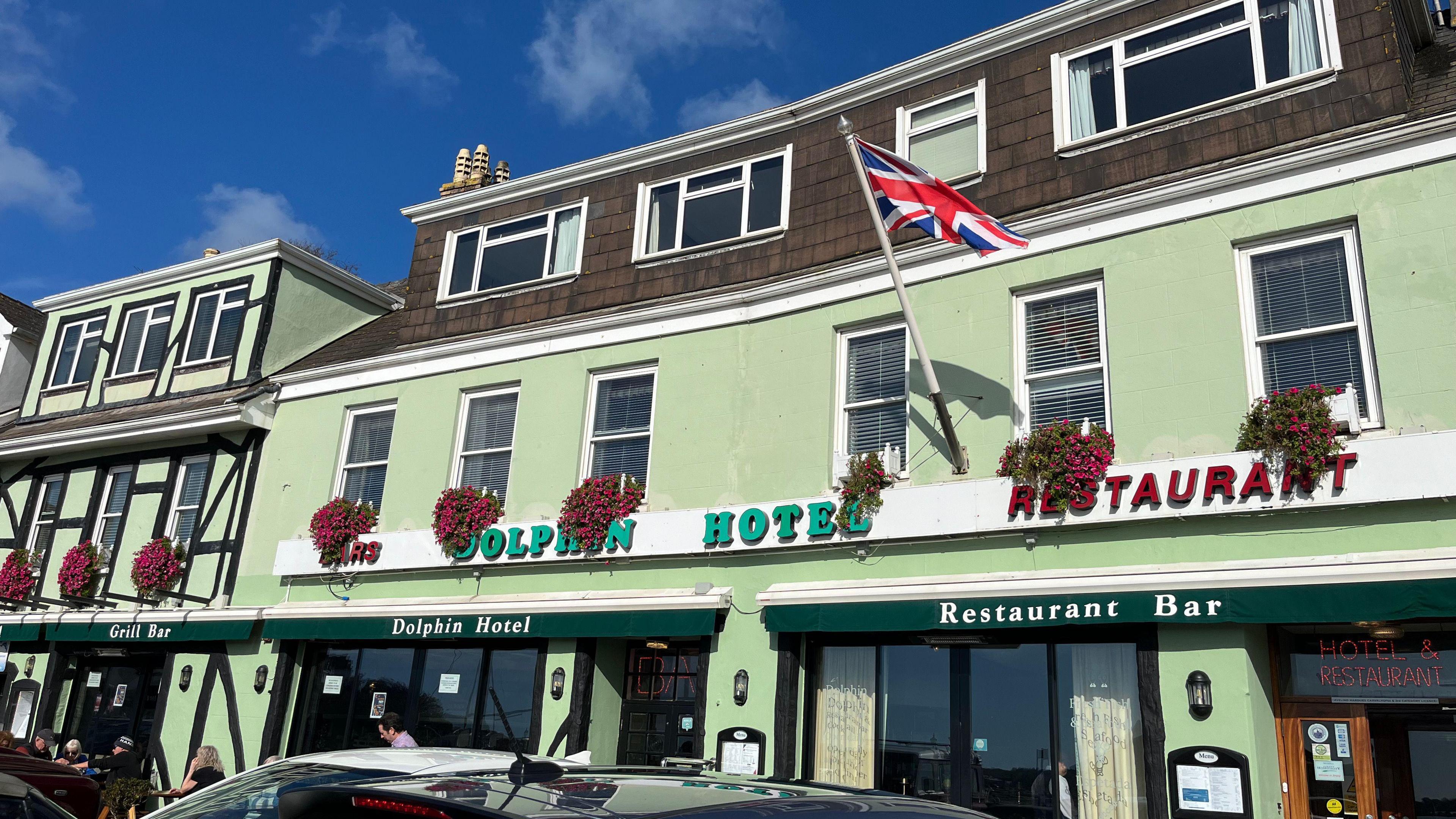 The Dolphin Hotel in Jersey which has green exterior walls and some cars parked outside it on a sunny but cloudy day. About half a dozen people are sat on benches outside the front of the building.