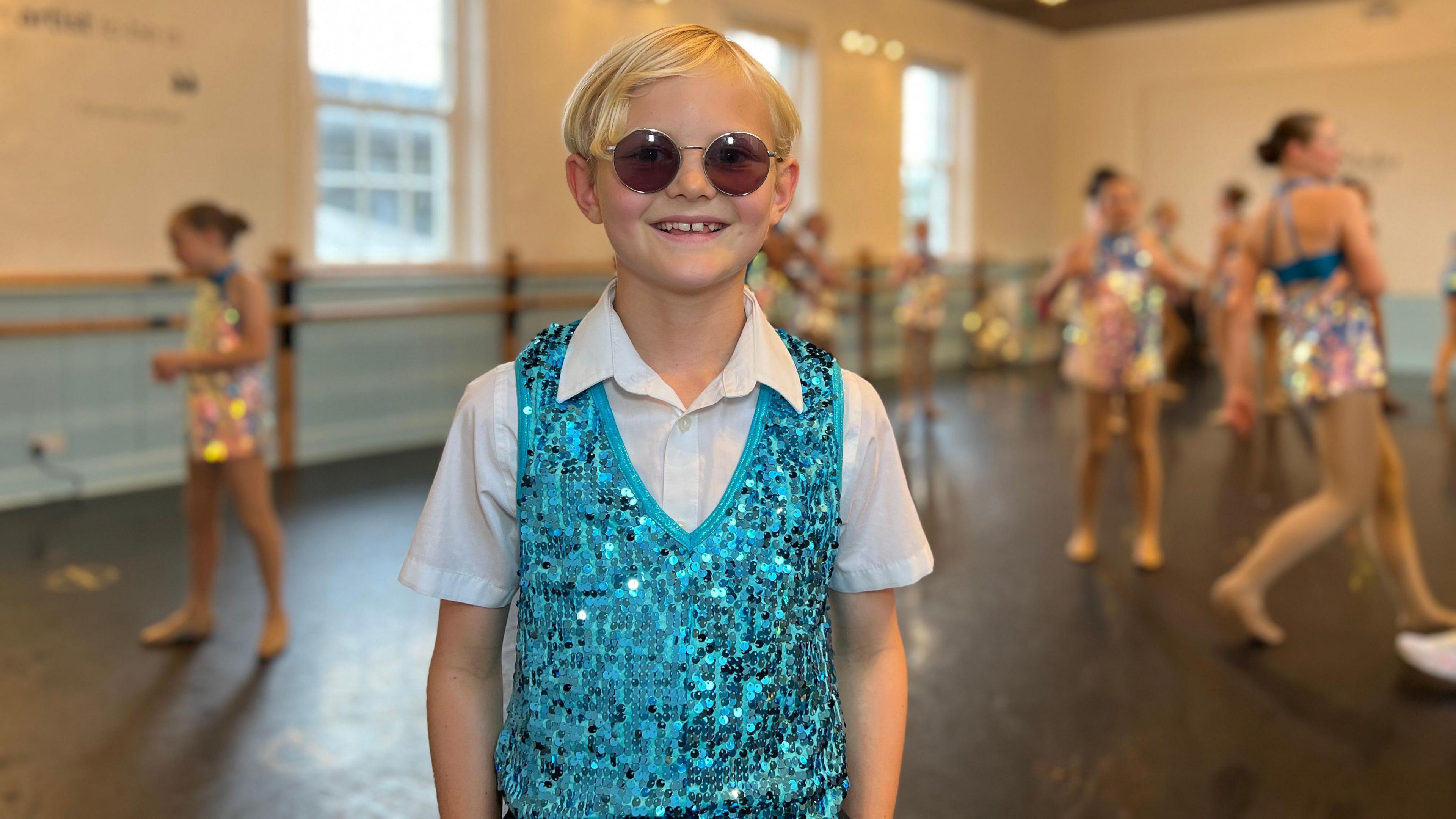 Henry wears a light blue sequin tank top, white T-shirt and sunglasses as he smiles at the camera