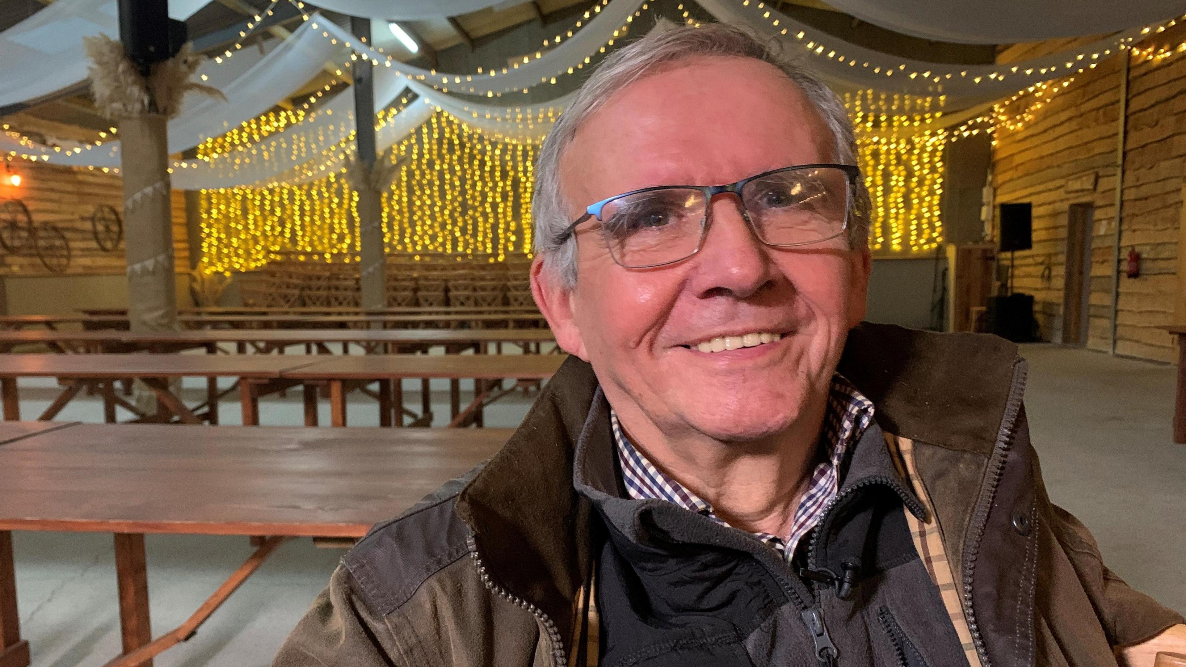 A man with grey hair and glasses, wearing a brown jacket, sat inside a wedding venue. It is a head and shoulders shot, and he smiles at the camera.