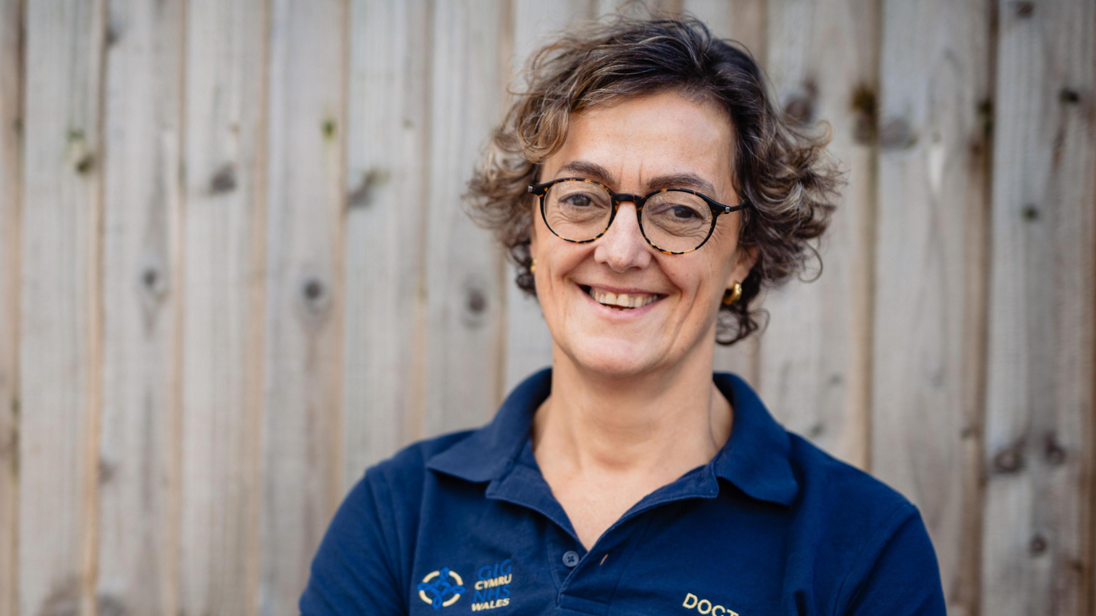 Dr Hilary Williams stood in front of a fence wearing a dark blue polo that says doctor and NHS Wales on it