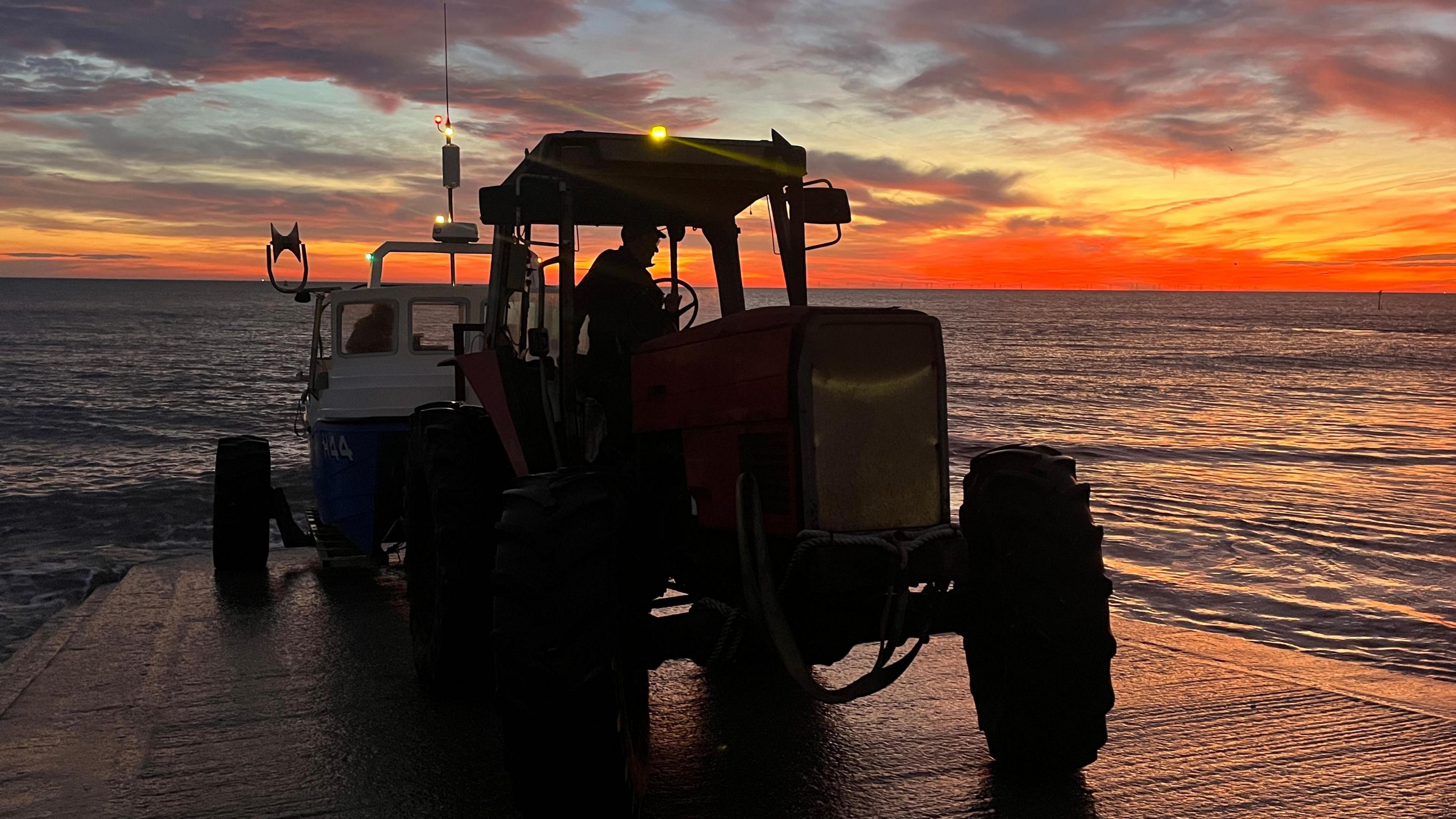 A tractor with a boat hitched on the concrete slipway at Hornsea, the sea is dark and calm and the sky is orange and red as the sun rises behind the boat.