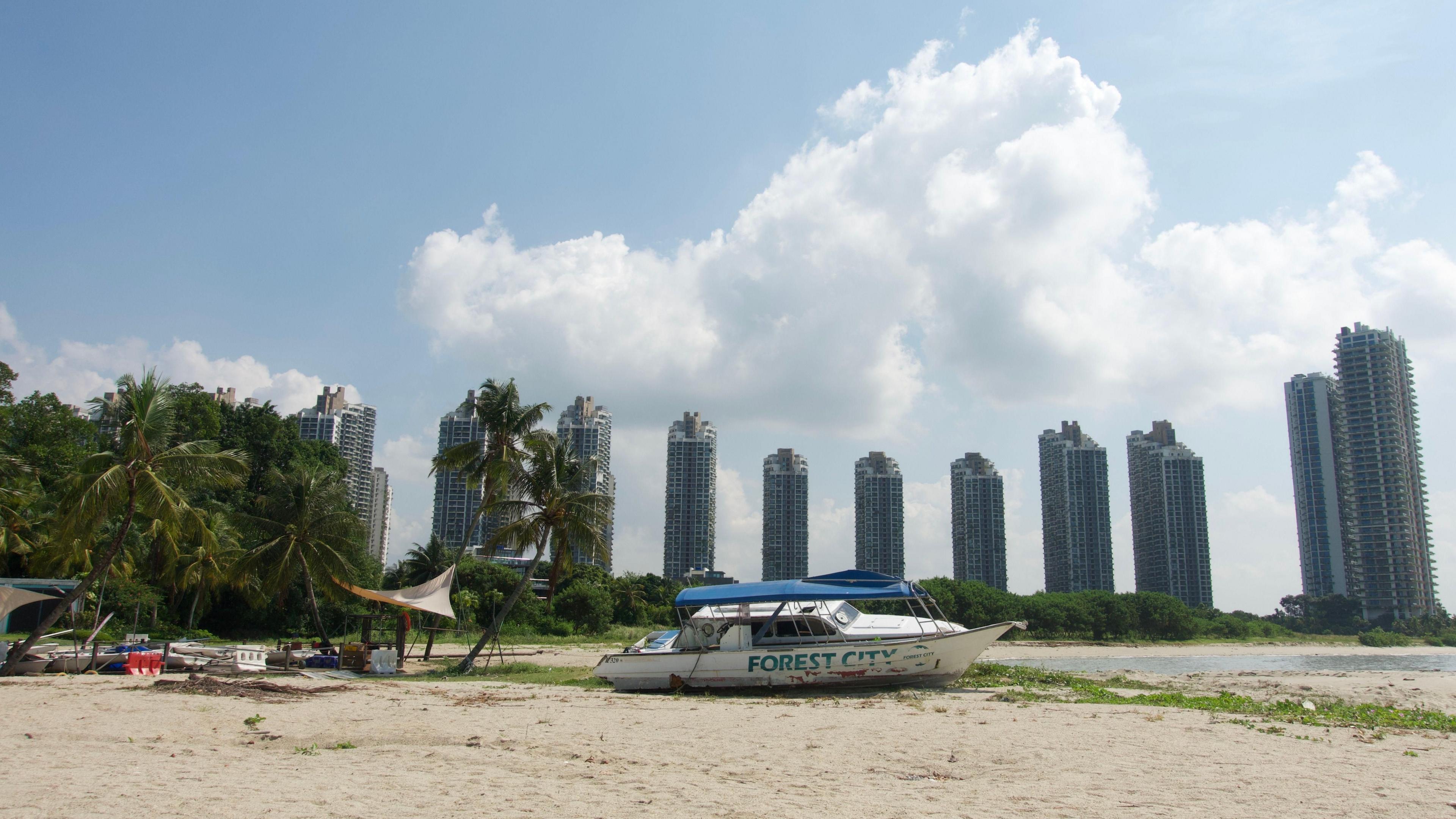 A dilapidated boat with the word 'Forest City' on its side sits on the sand of a beach, next to a pile of random debris and palm trees, with high-rise buildings in the distant background