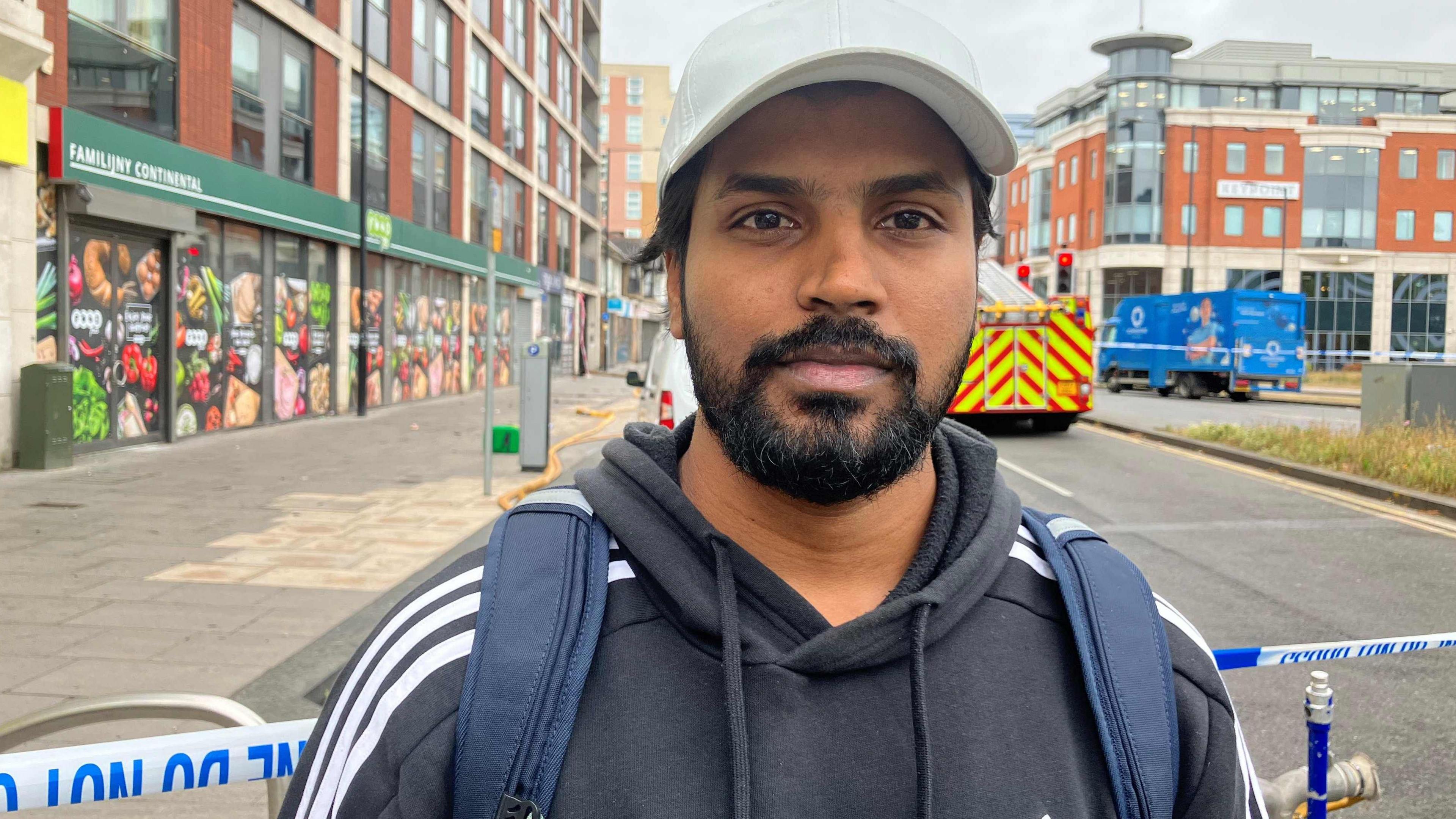 Rahul Shelke looking straight down the camera lens, has facial hair and is wearing a hoody in navy with white stripes and a white basketball cap. He is standing in front of a police cordon tape with the block of flats in the background (which he was evacuated from) due to a building fire. 