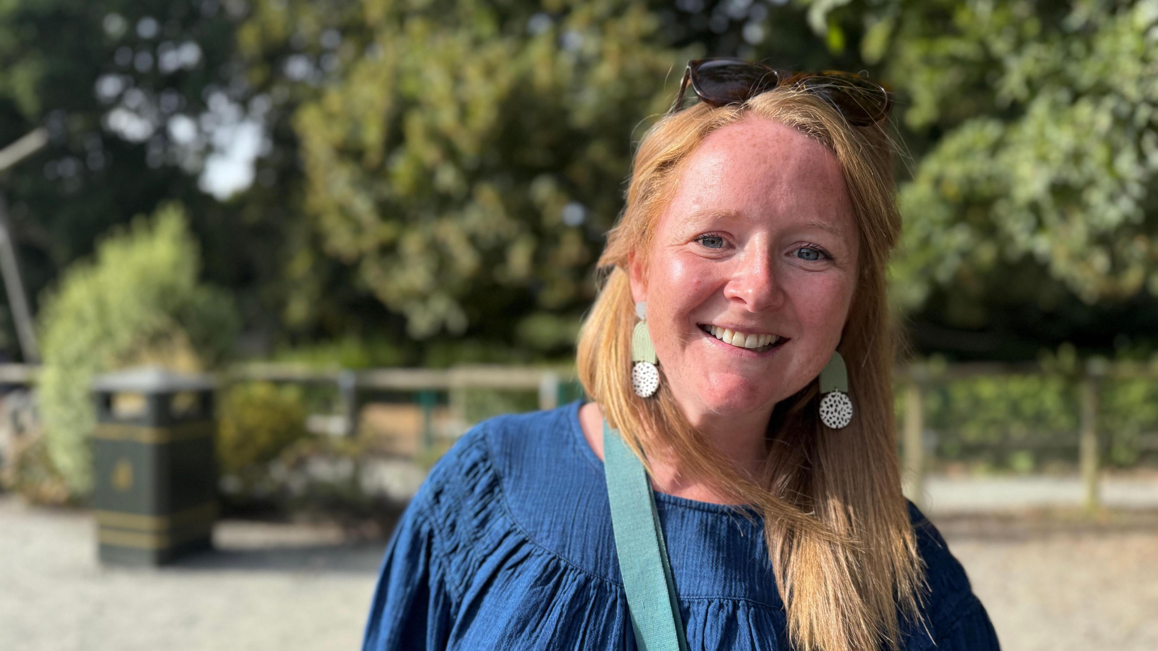 Woman in her 30's with long ginger hair. she is in a denim blue top and big earrings