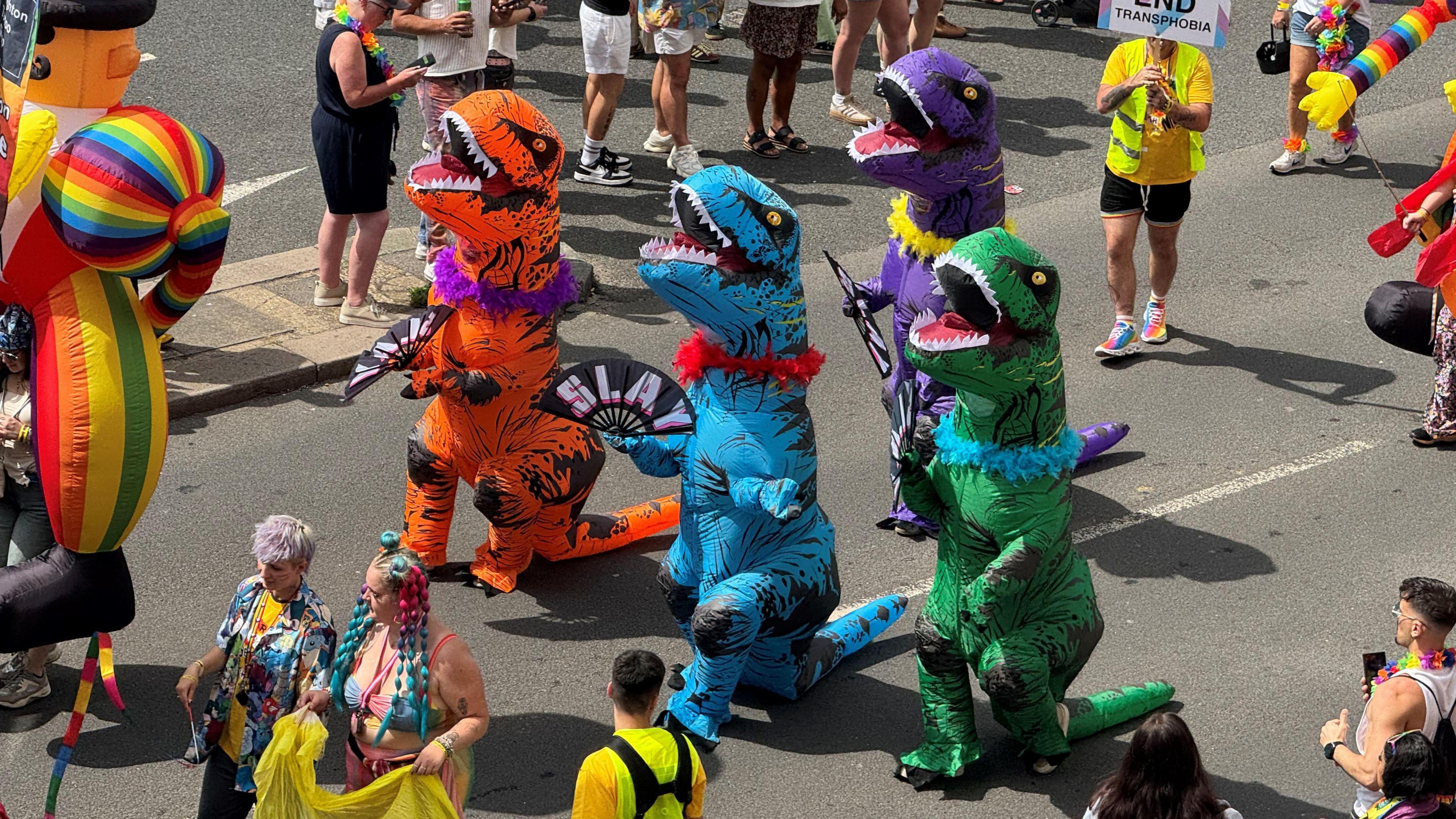 A group of people wearing multi-coloured dinosaur costumes. They are holding fabric fans with the world "Slay" on them.