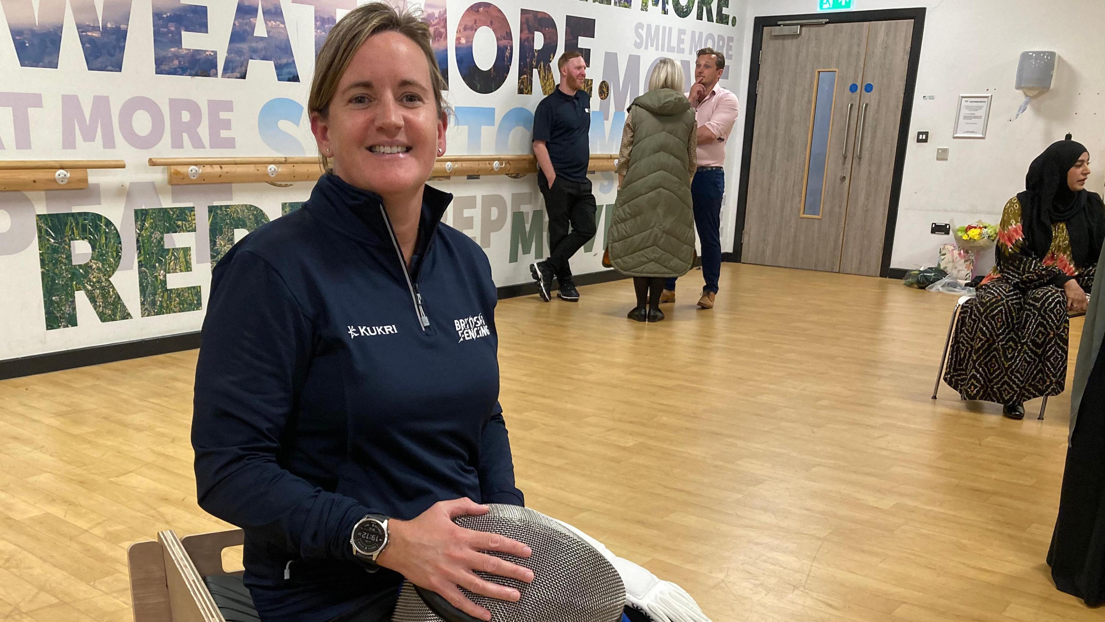 Virginia Bailey sits on a sword seat and holds a fencing mask on her knee. She is wearing blue sports kit and has her hair tied back.