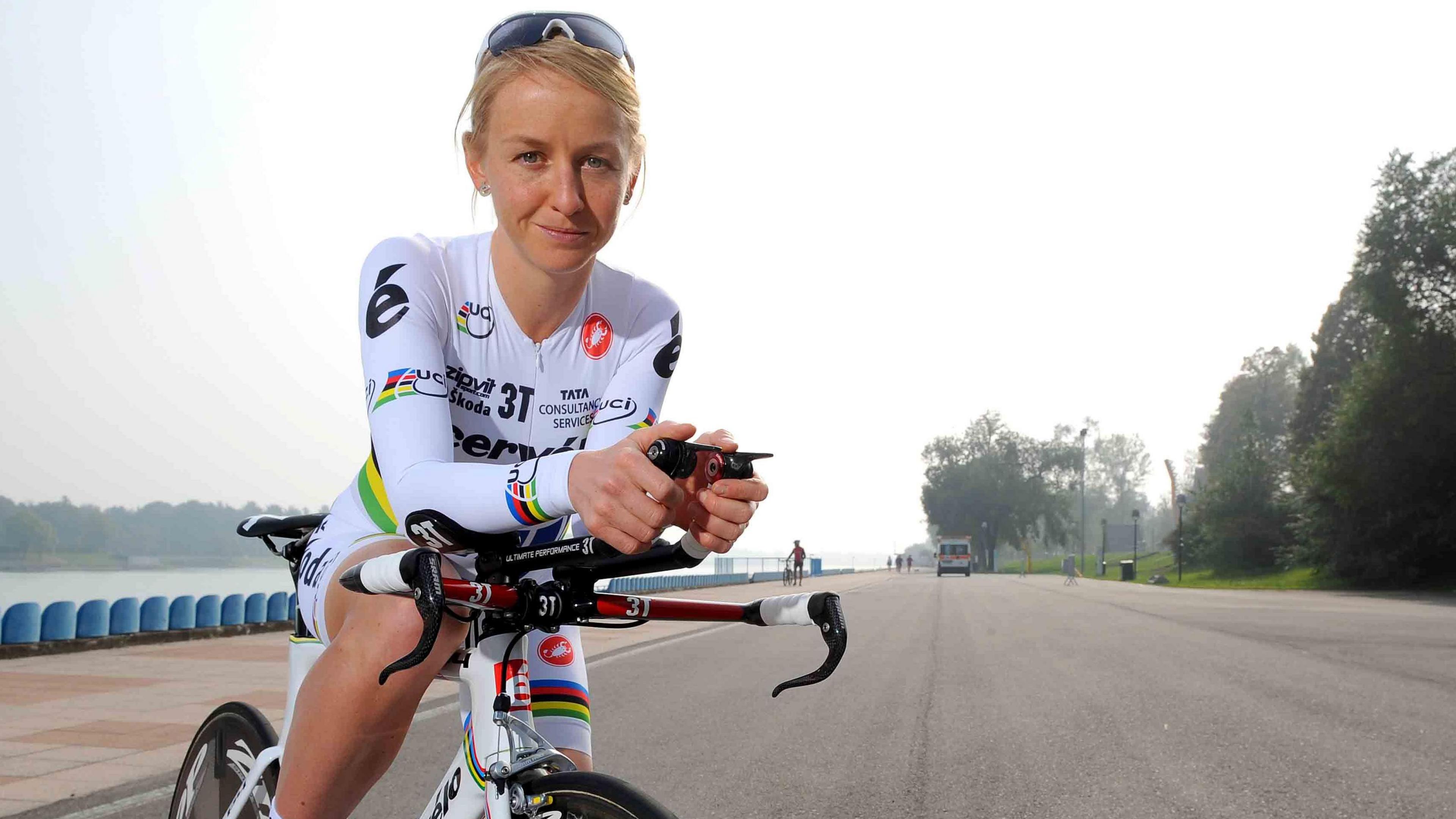 Emma Pooley is looking at the camera while posing for a shot on a bicycle. She has blond hair and sunglasses are balanced on her head. She is wearing a white cycling suit. There is a road behind her and in the distance are people and a van