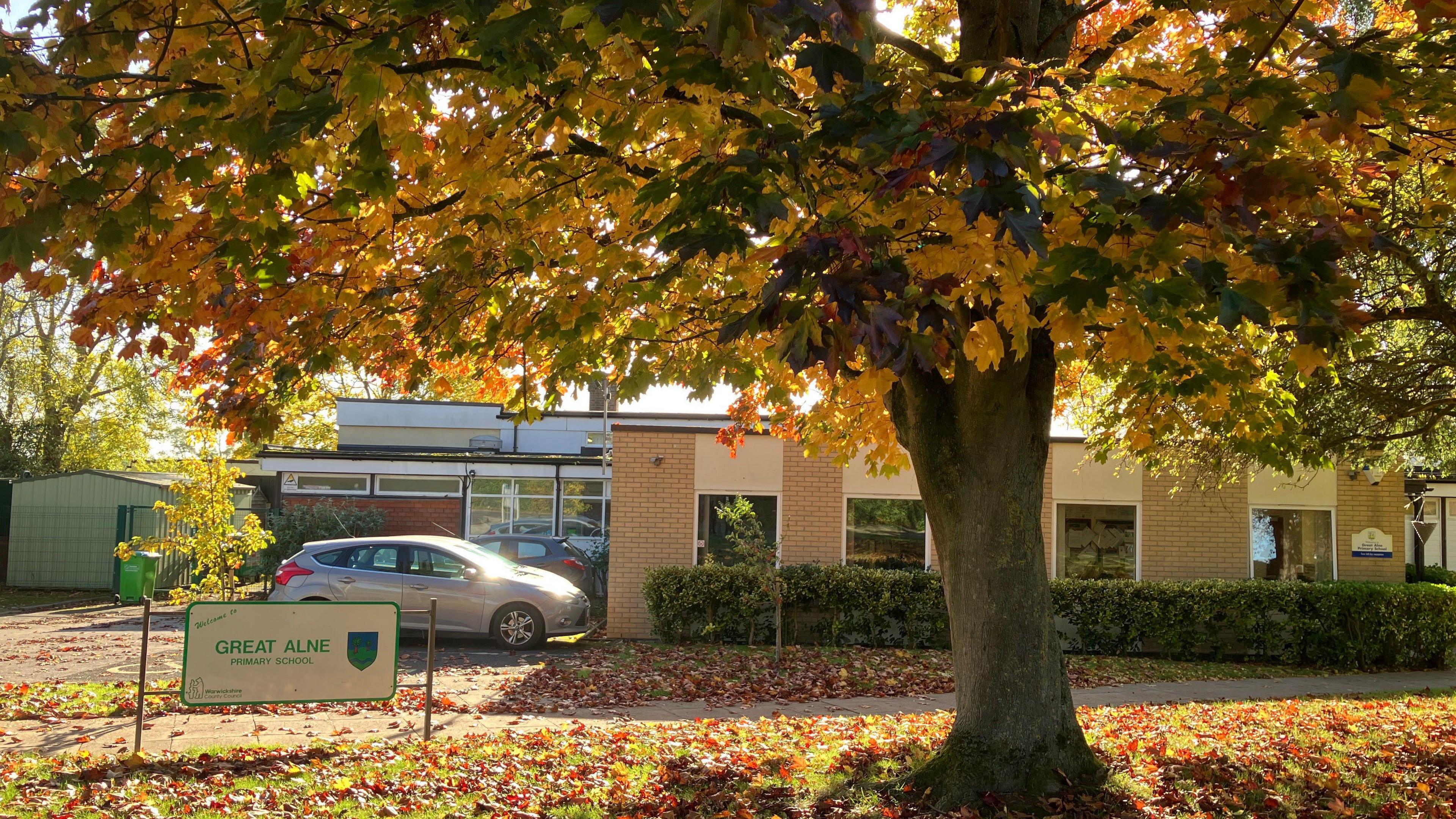 The school building is seen with autumn leaves around it. There is a school sign under a tree and cars are parked outside.
