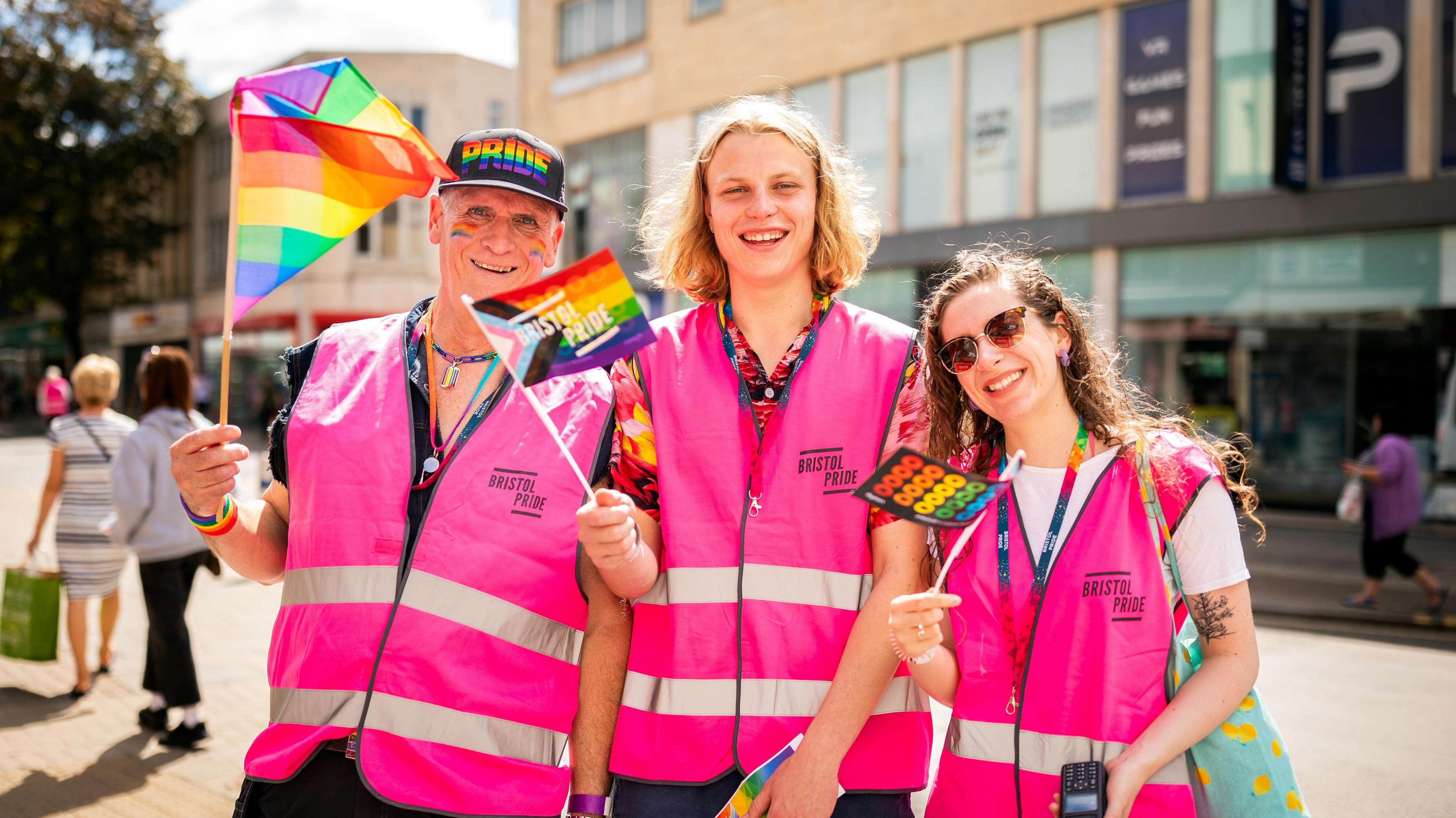 Three volunteers wearing pink high-vis vests and holding different rainbow flags. 