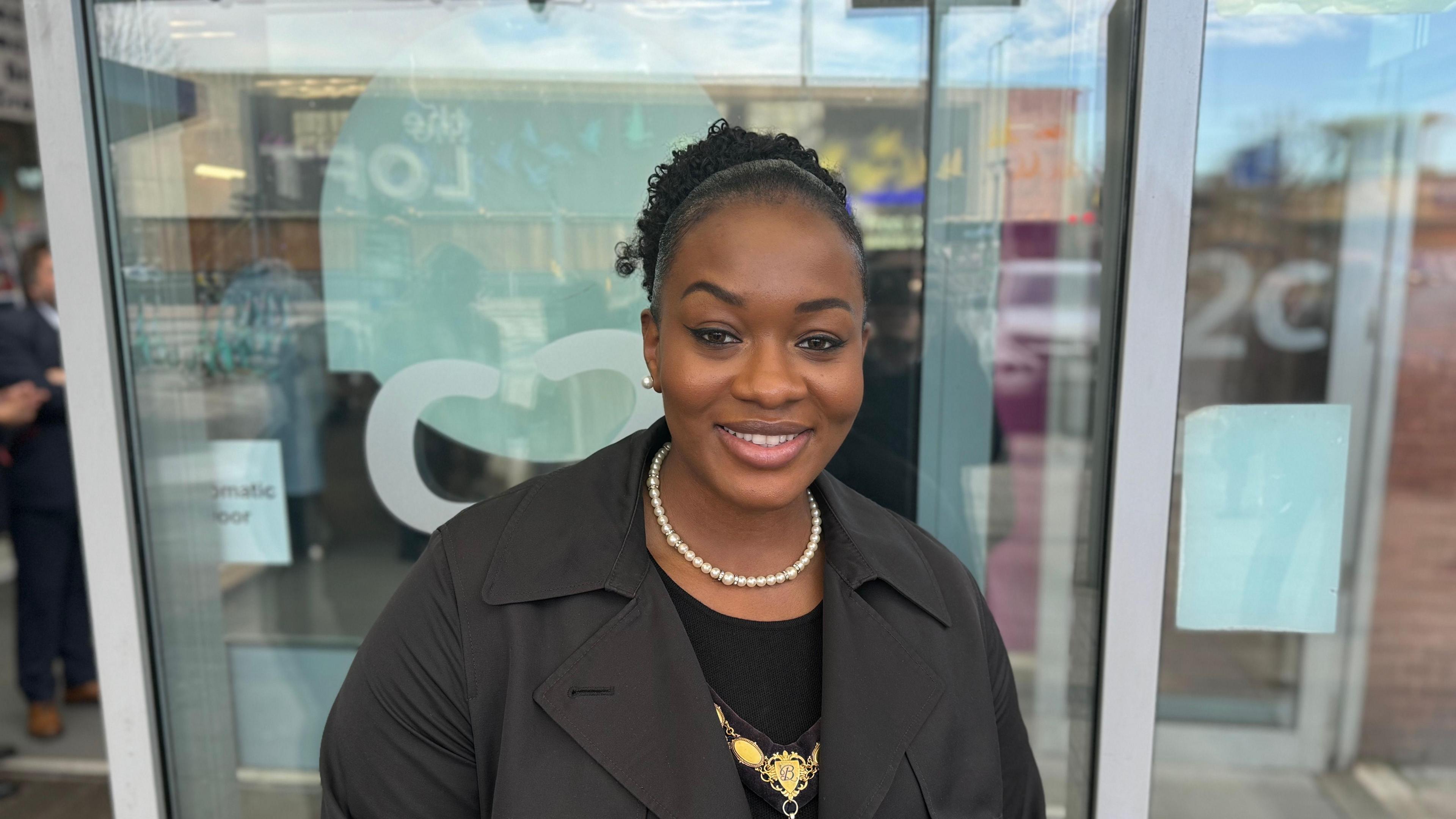 A black woman standing in front of a glass door smiling. She is wearing a black coat and a white beaded neckless. Behind her is the transparent automatic sliding doors of the station