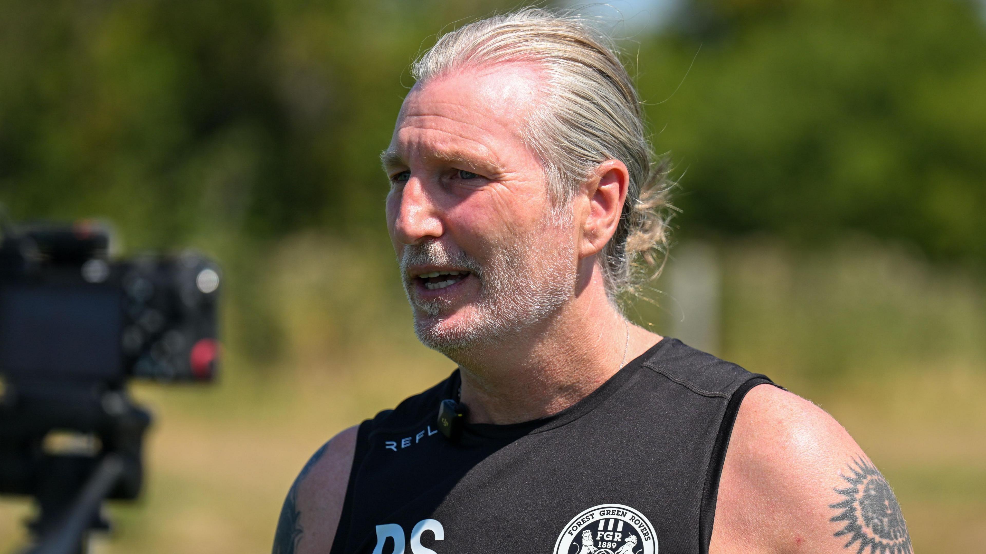 Forest Green Rovers boss Robbie Savage wearing a black vest with his initials on, and the club logo on, with his left arm covered in the sun, revealing his tattoos.