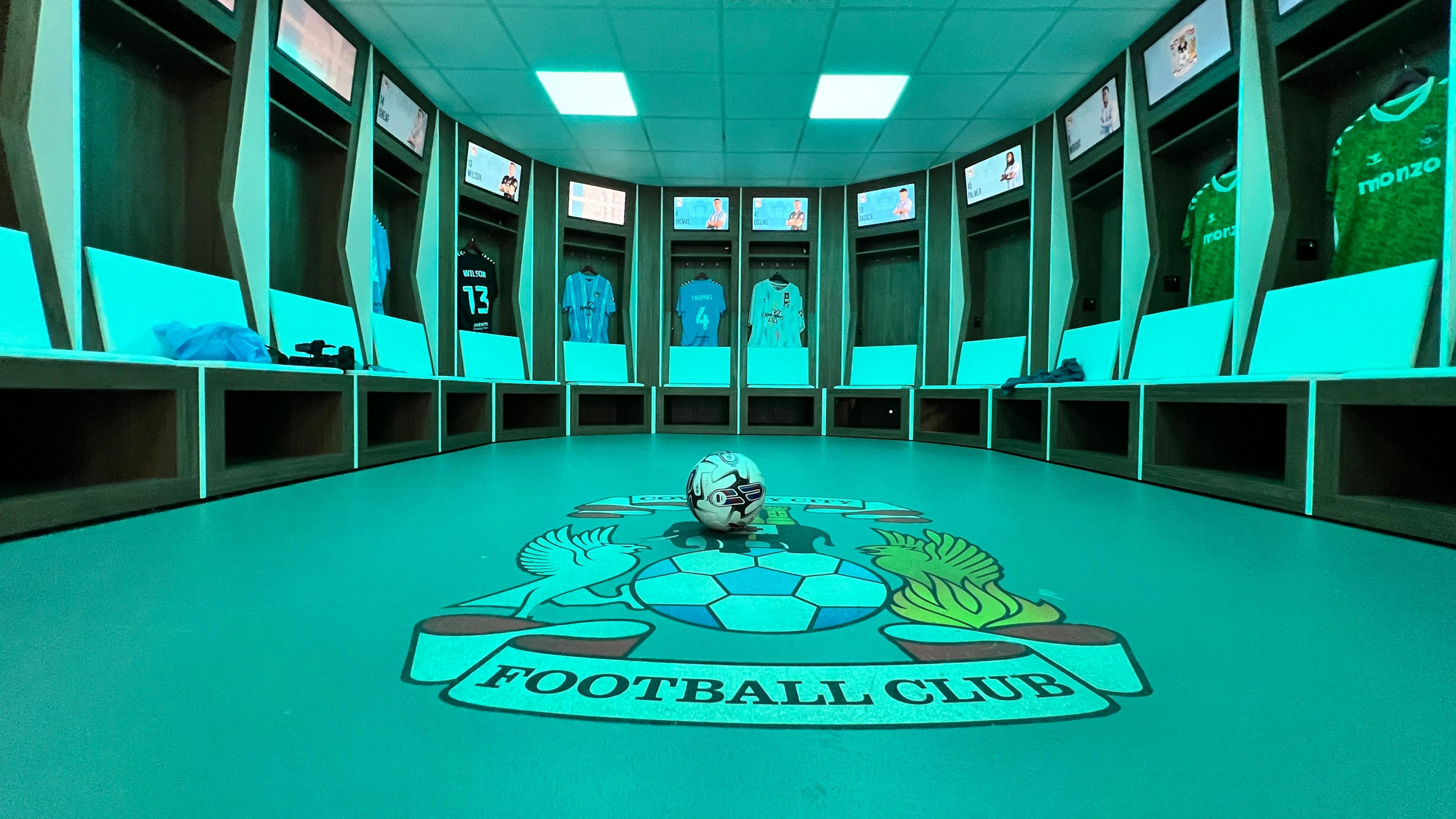 An empty football stadium dressing room, the floor has the Coventry City logo on it, and the whole room is lit in a blue tone