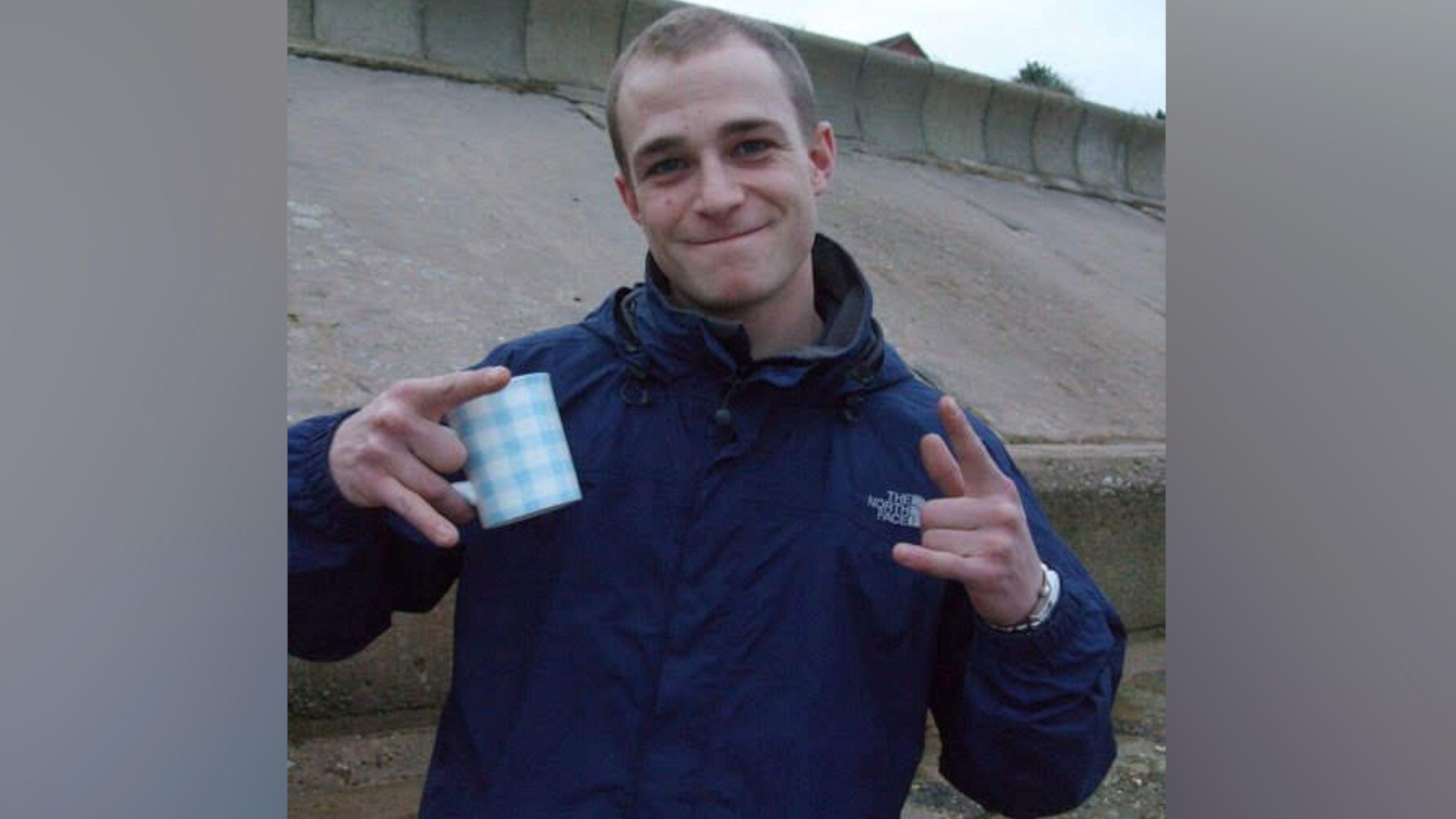 Terry White stands in front of a grey concrete sloping sea wall. He has short cropped brown hair and is smiling at the camera. He is wearing a navy blue jacket with a white The North Face label on the left of his chest, and he is holding a white and blue mug in his right hand.
