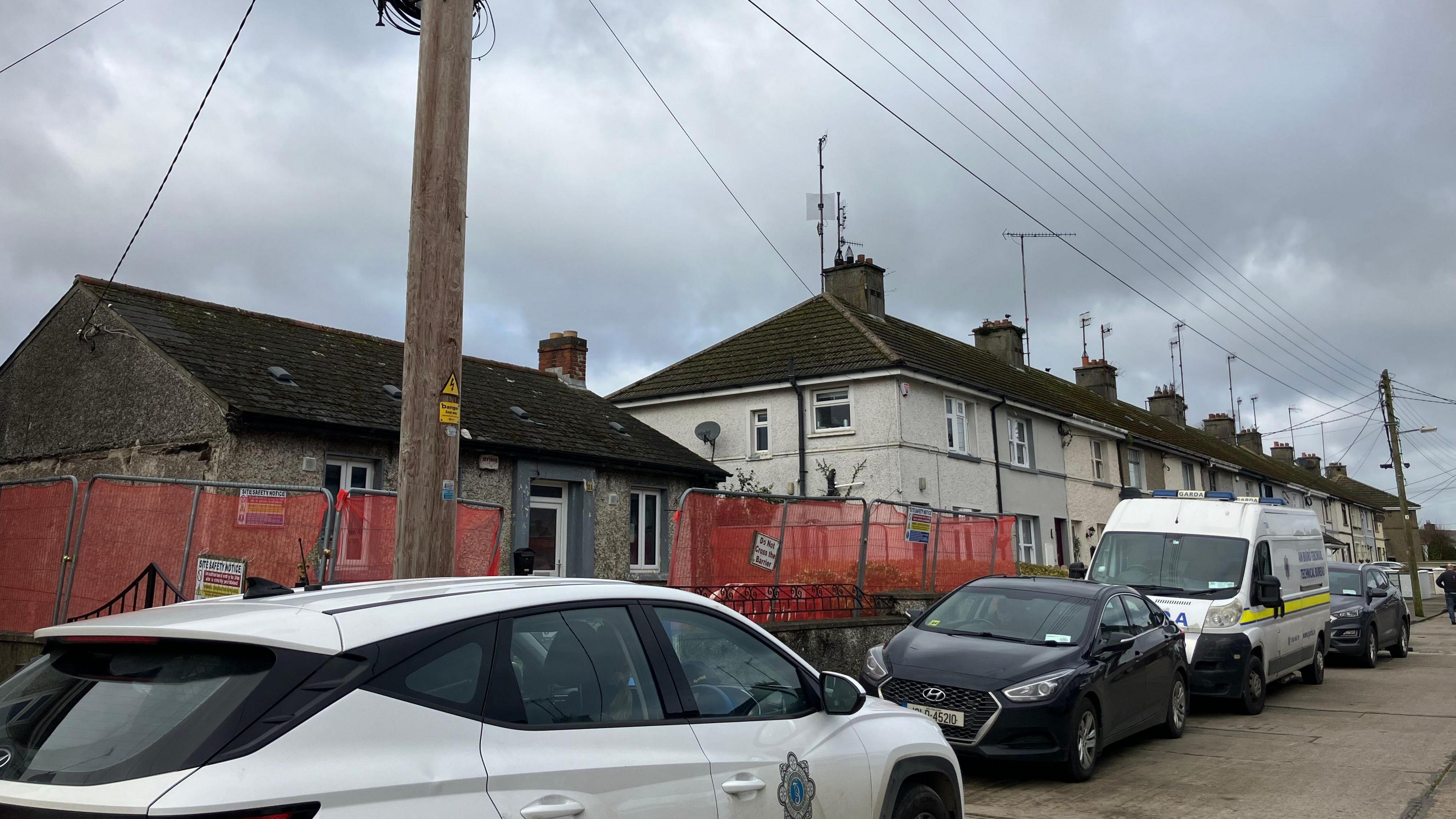 A row of houses in Drogheda, the closest house seems to be abandoned with red railings surrounding it
There are a row of cars parked, among them a white Garda van and Garda car