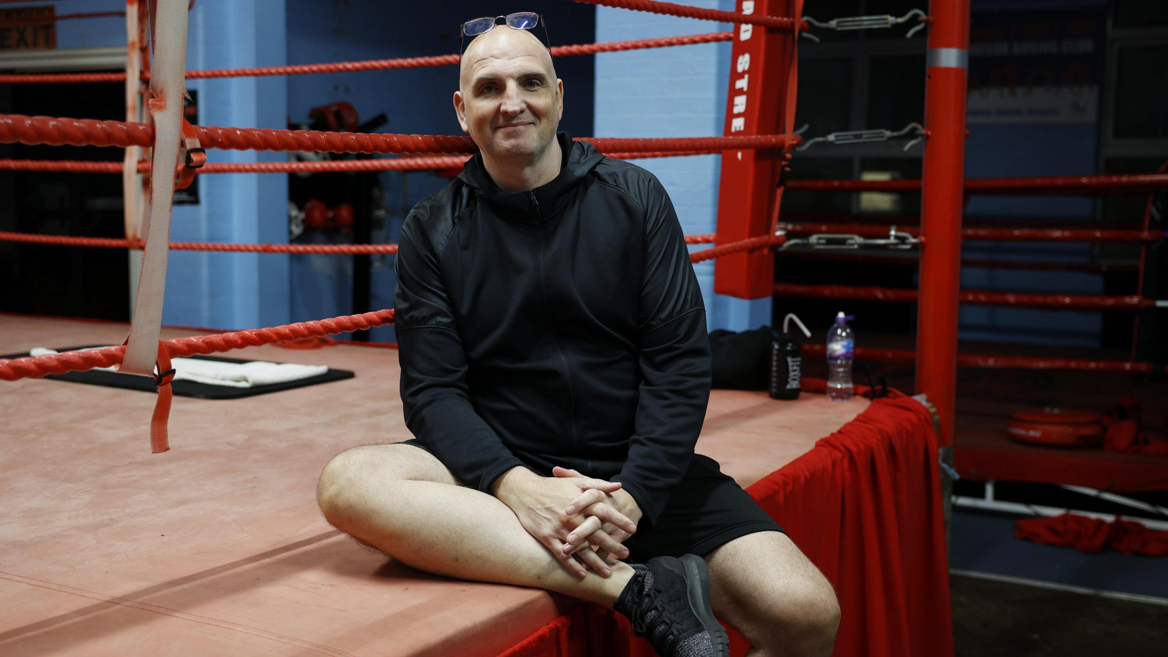 Ray McCallum in a black hoodie, black shorts, black trainers and glasses on his head. He is sat on the side of a red boxing ring.