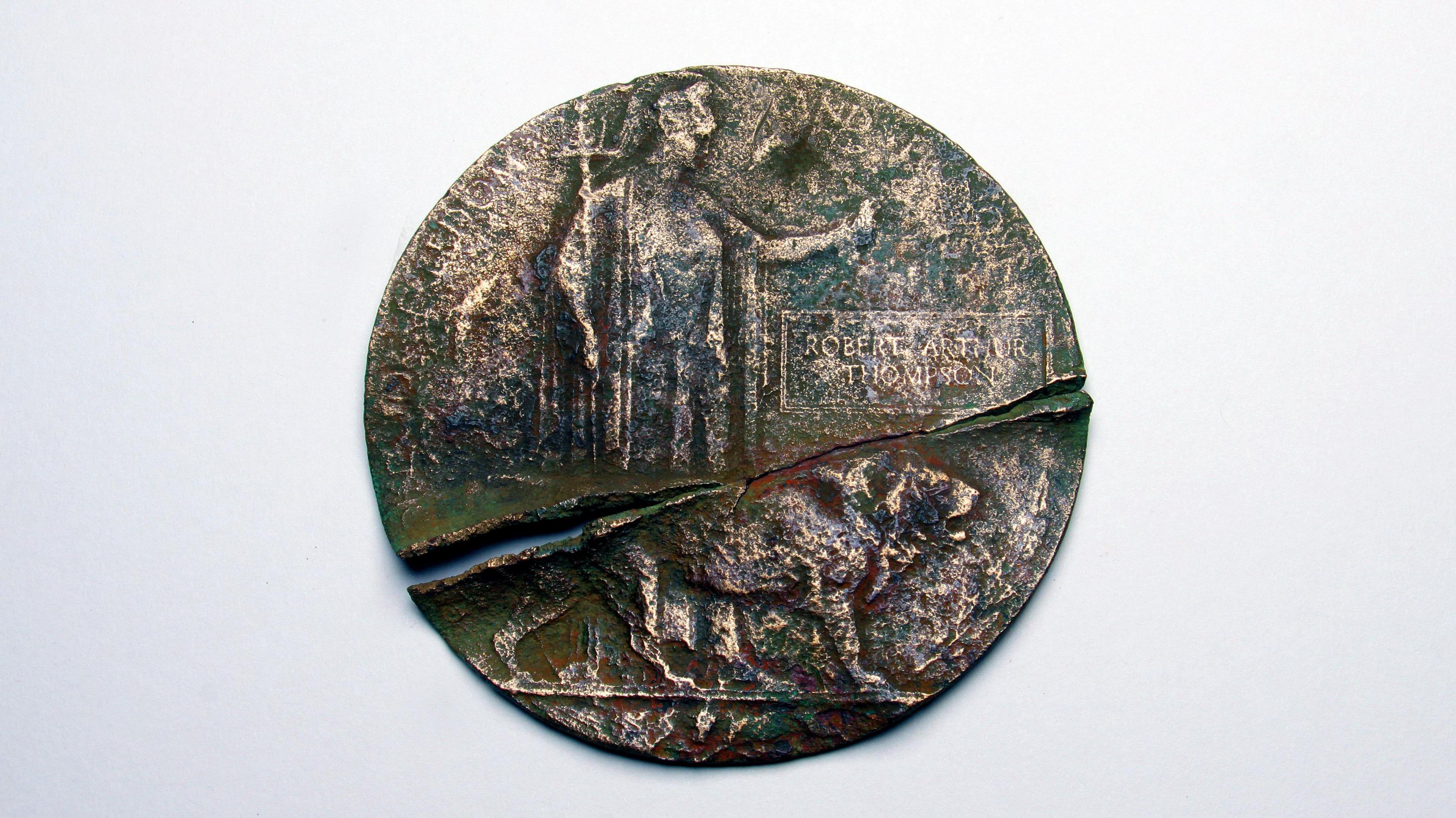 An old bronze plaque. It is circular and split in two. On it is the figure of Britannia, a helmeted female warrior holding a trident and shield, with a lion at her feet. The name of Robert Arthur Thompson is engraved on the plaque within a rectangular border, between Britainnia's outstretched left arm and the lion's head.