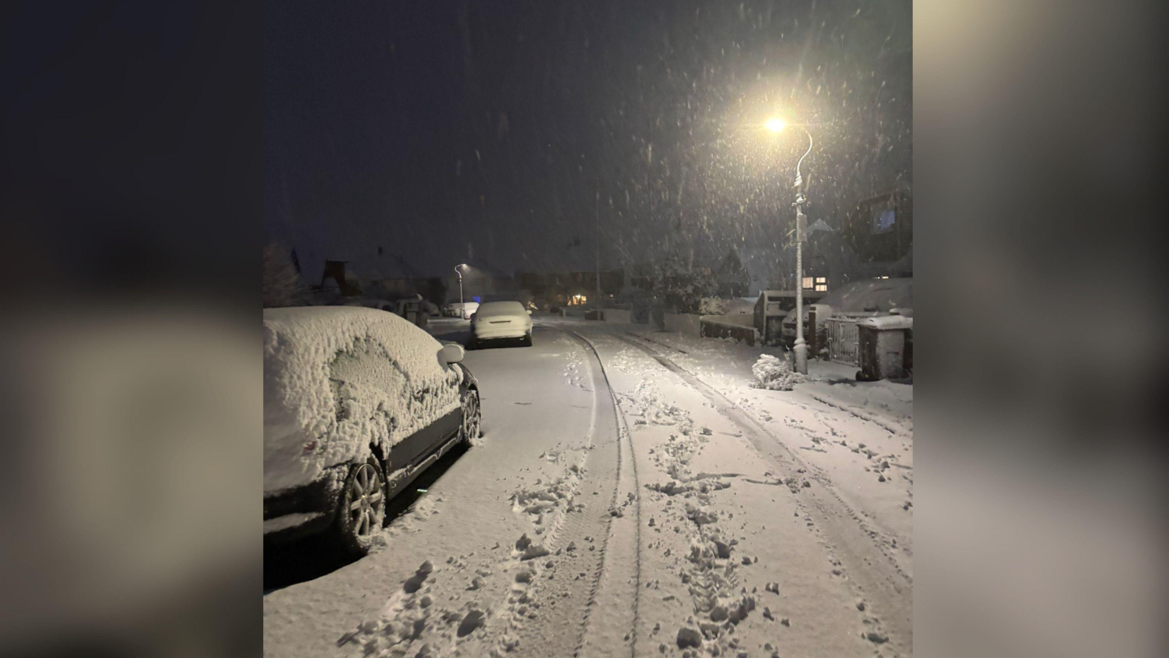 A road covered in white snow. There is a street light on the left and a number of cars covered in snow on the right. 