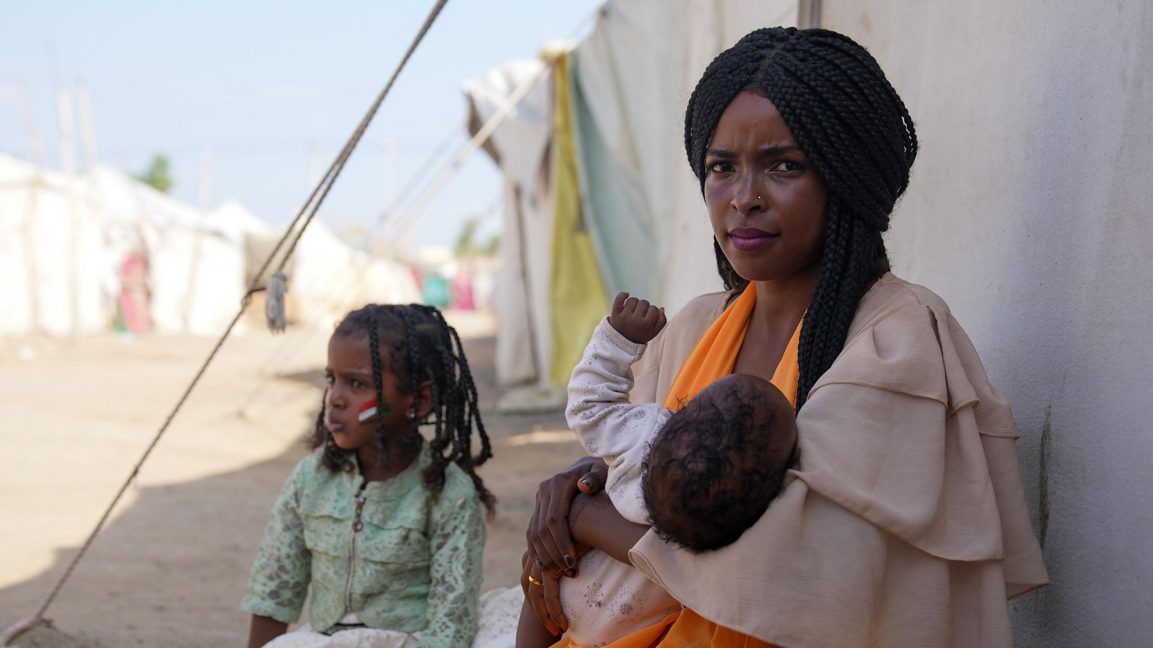 A woman called Omnia sits outside a tent in Sudan holding her baby, with a second child sat next to her