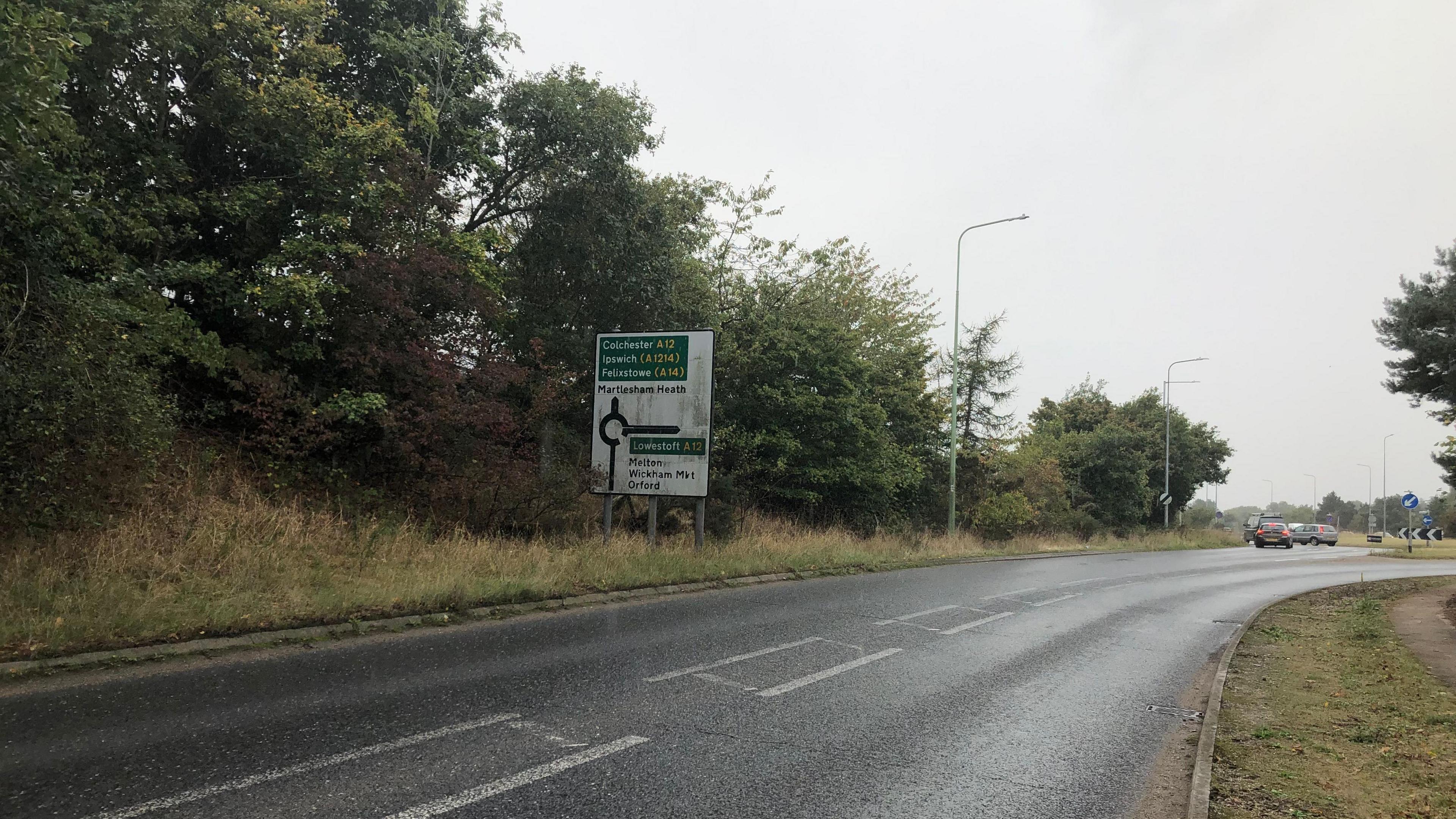 Part of the A12 through Suffolk. It shows a road leading up to a roundabout. Cars can be seen in the distance making use of the roundabout. There is a road sign and trees and bushes line the road.