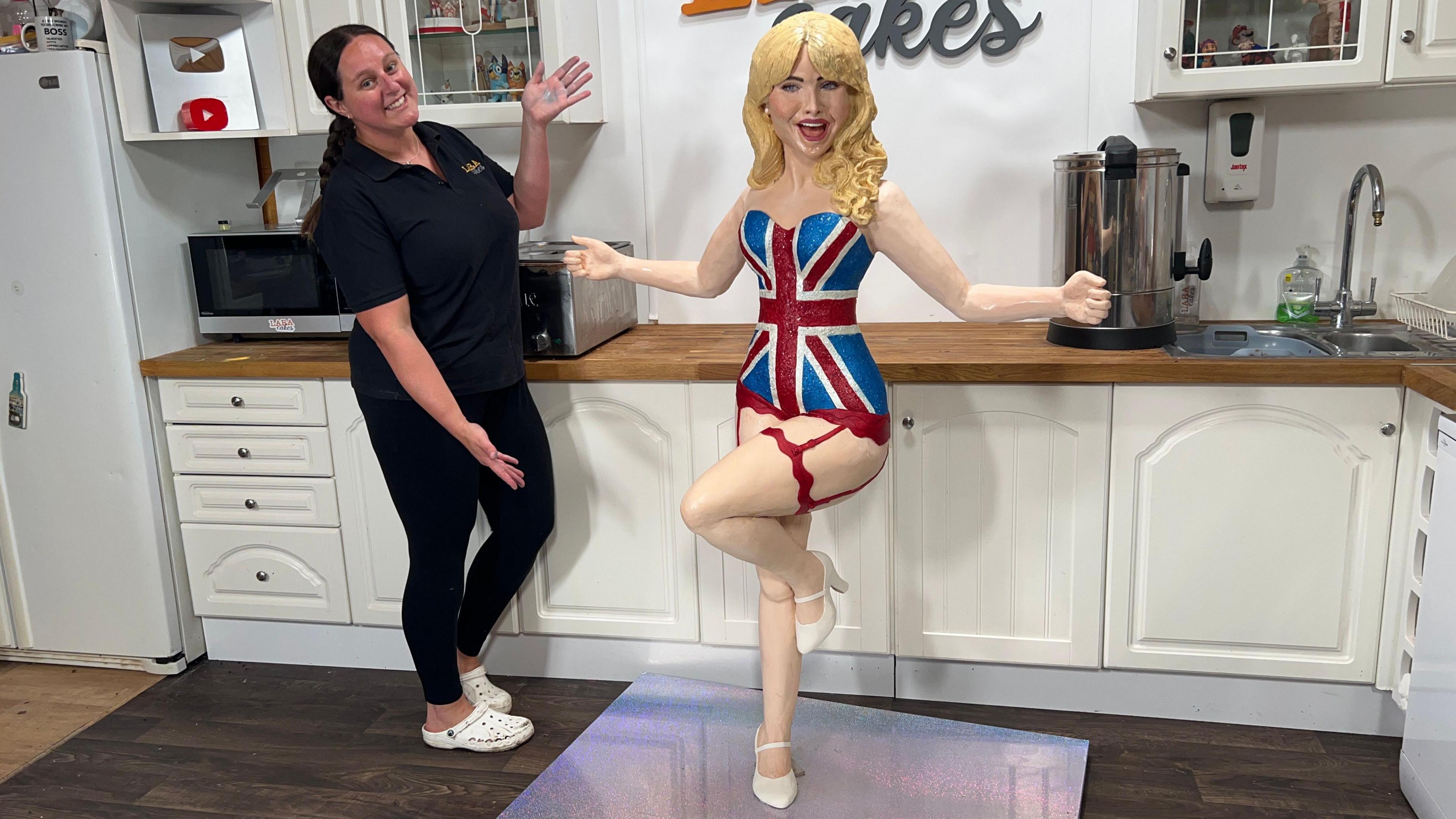 A woman dressed all in black with white croc shoes is standing next to a 5ft cake designed to look like Sabrina Carpenter