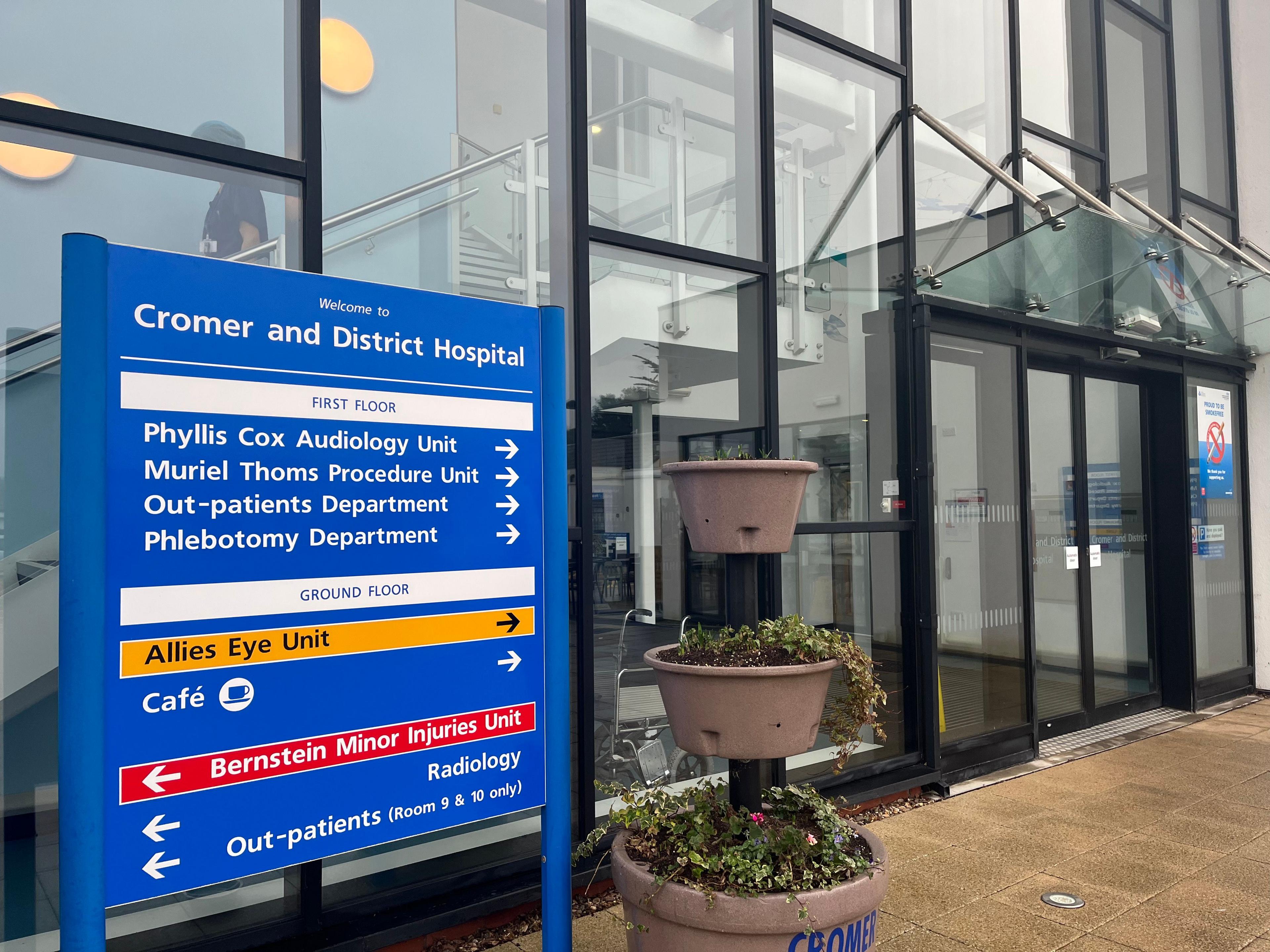 External view of Cromer Hospital. 
It is a glass building with a blue sign outside with a list of all the different departments and services on offer with arrows showing where visitors should go. 