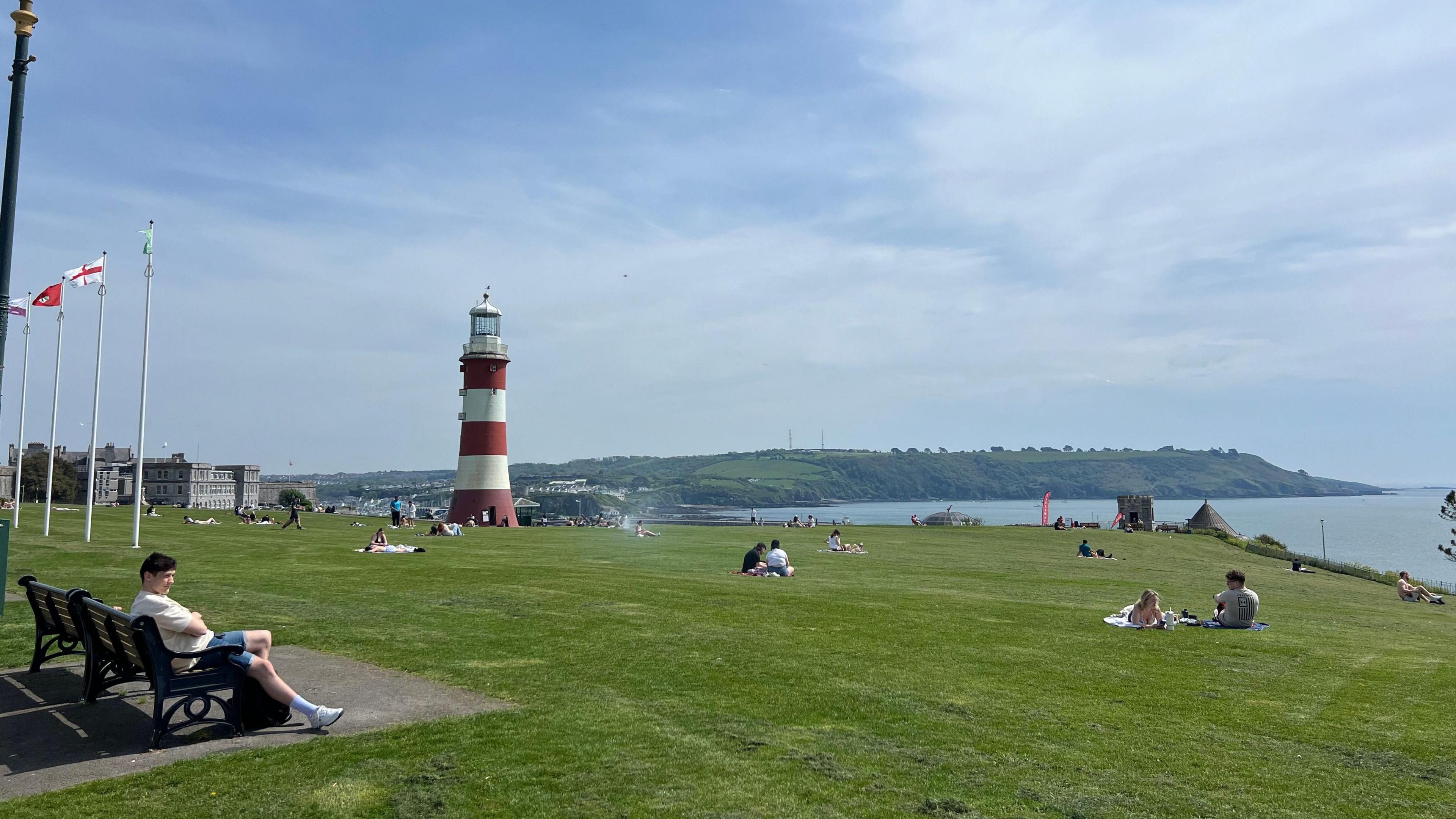 A general view of Plymouth Hoe. There is a large grass area, with a lighthouse in the distance. People are sat in groups on the grass and in the distance is the sea and land.