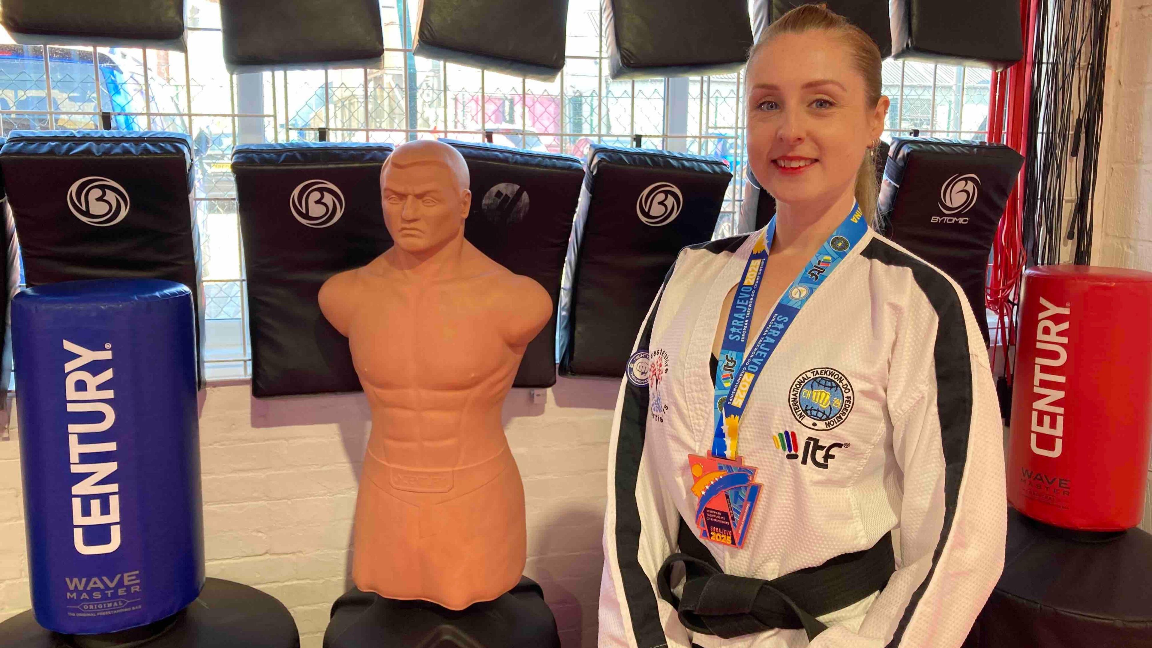 A woman wearing a white jacket, trousers and a black belt, along with a lanyard, stands in a martial arts studio. Various equipment, including punching bags, cushions and a dummy can be seen behind her.