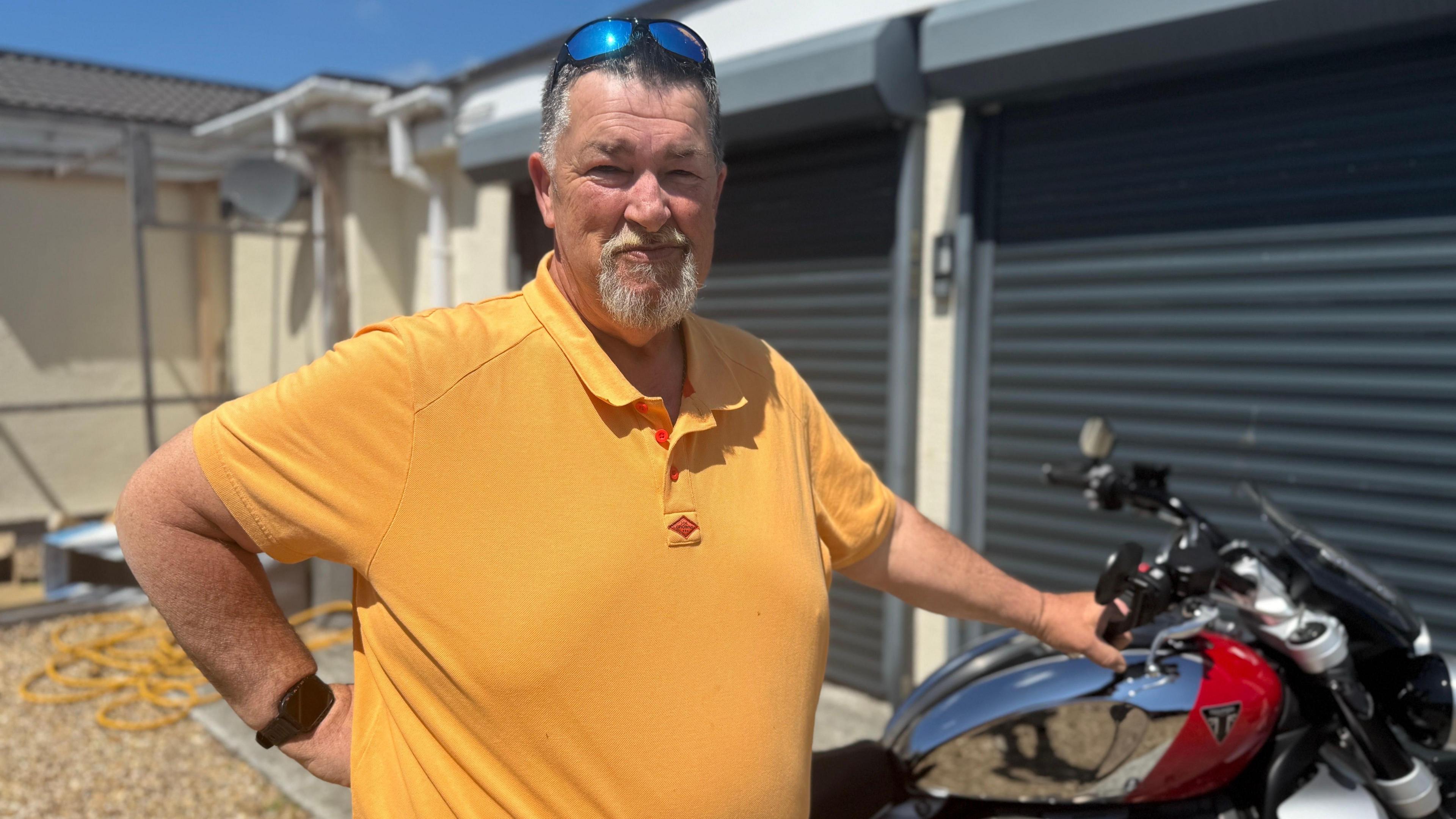 A man with short dark grey hair and a white goatee beard looking at the camera. He is wearing a mustard yellow polo and has sunglasses resting on his head. You can see a red and silver motorbike in the background
