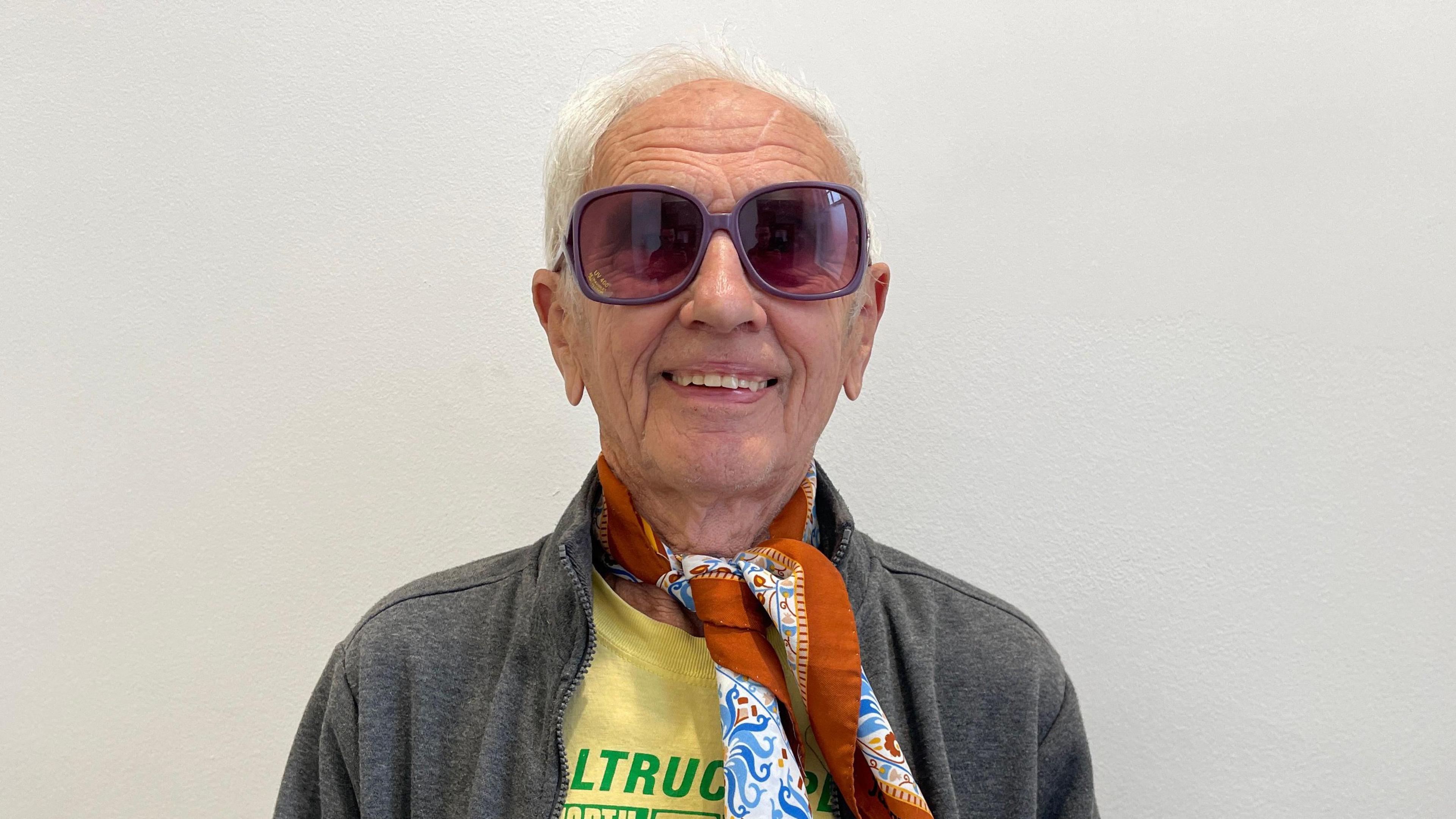 Bob Emmerson wearing dark eyeglasses and standing against a white wall looks directly at the camera.