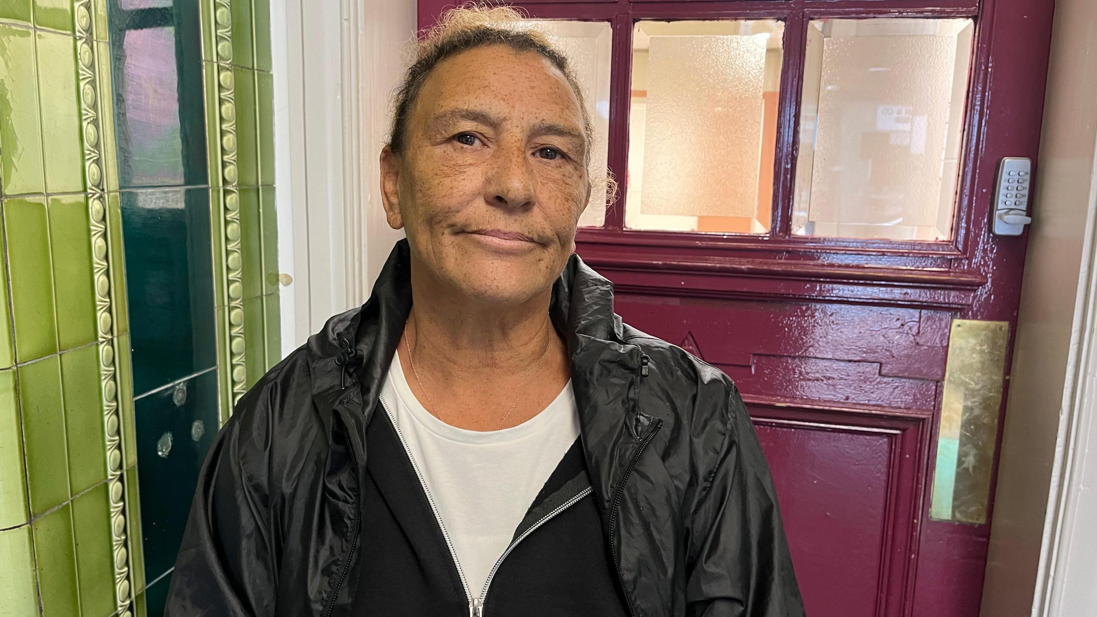 Lesley Thieme, 65, who has brown curly hair tied back and is wearing a black raincoat over a black cardigan and a white t-shirt, looks into the camera with a serious expression.