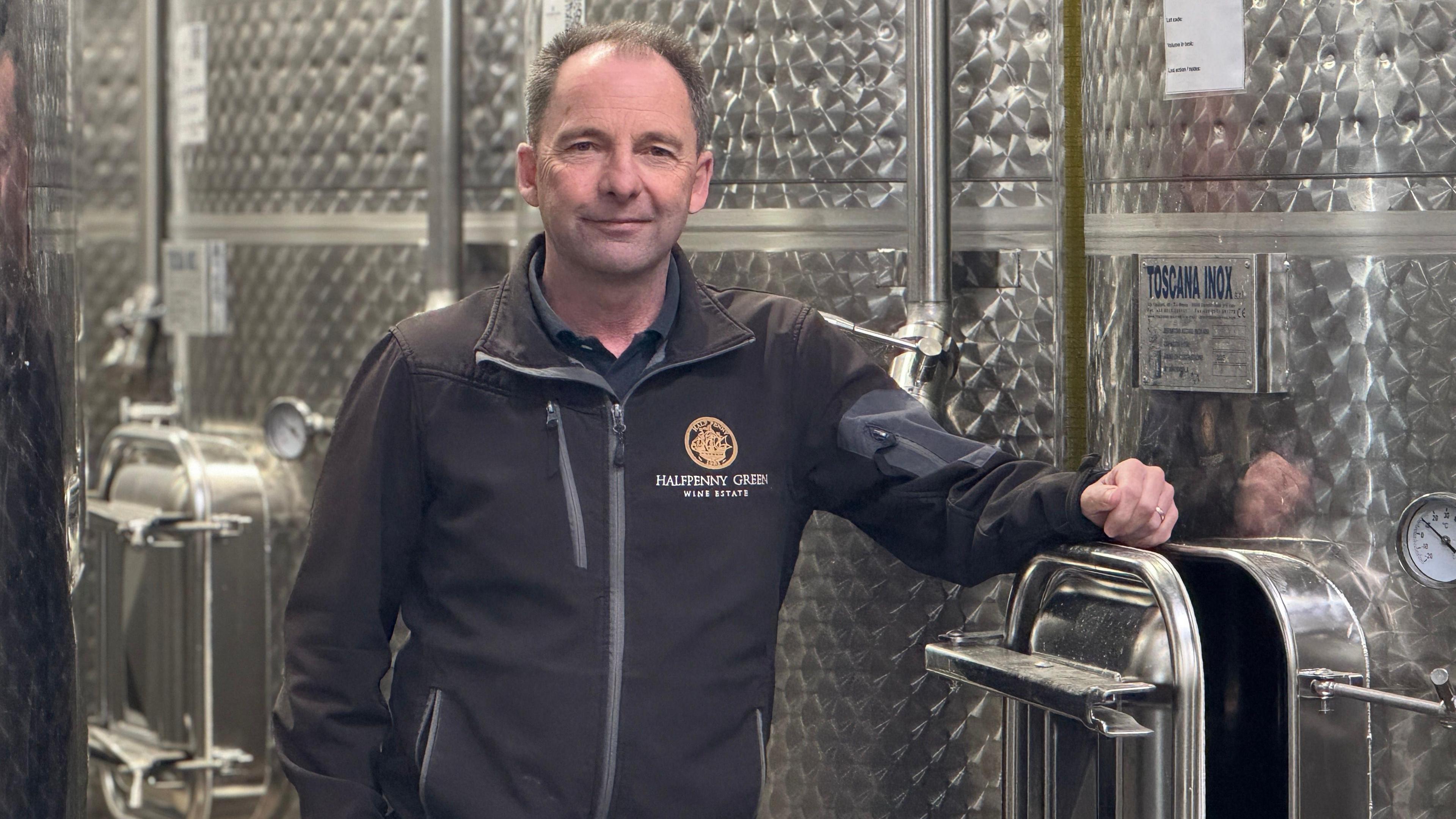 A man stands next to stainless steel containers and tanks.