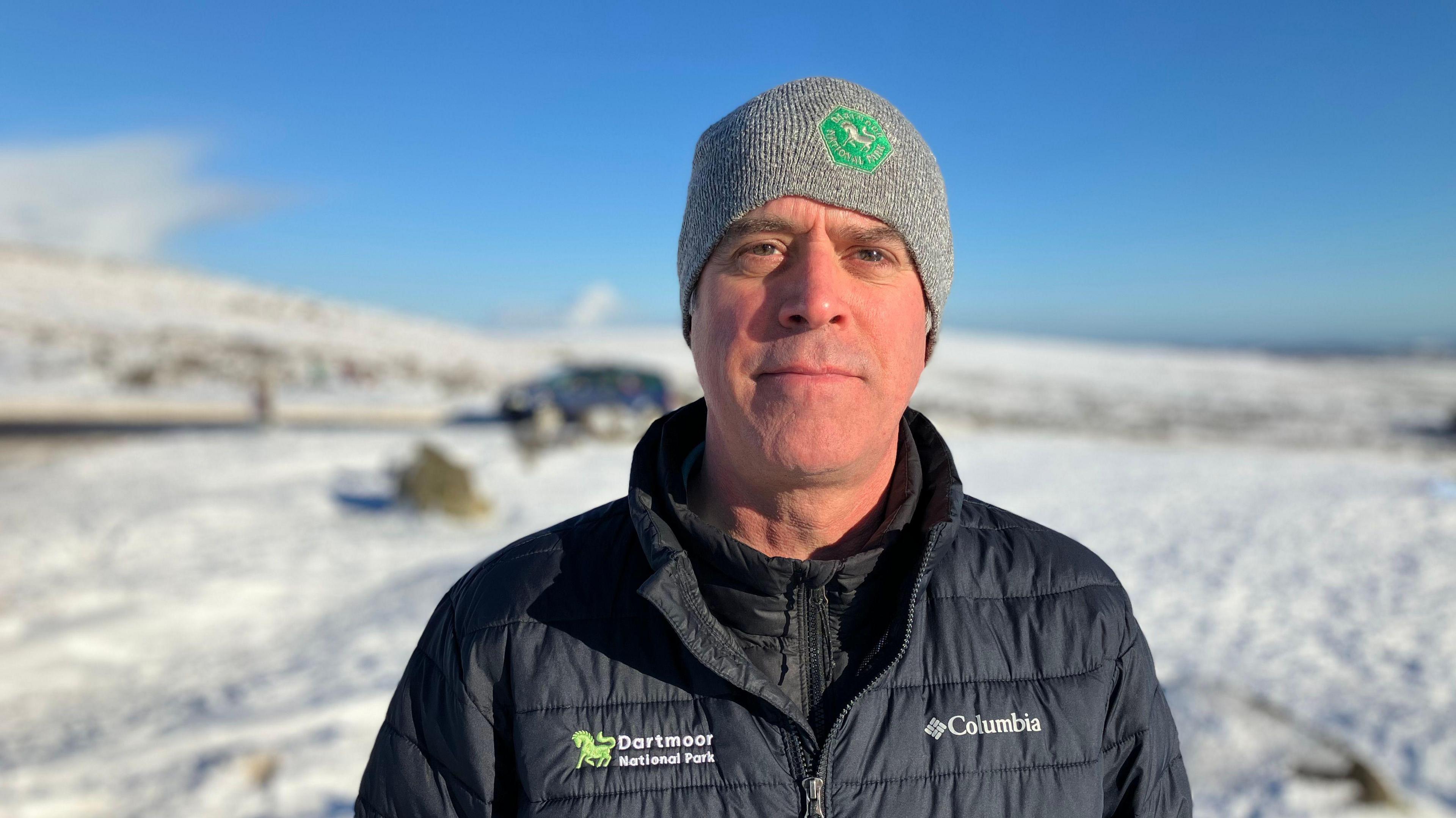 A man stands in a snow covered landscape. There is moorland in the background. He is wearing a hat and winter coat. 