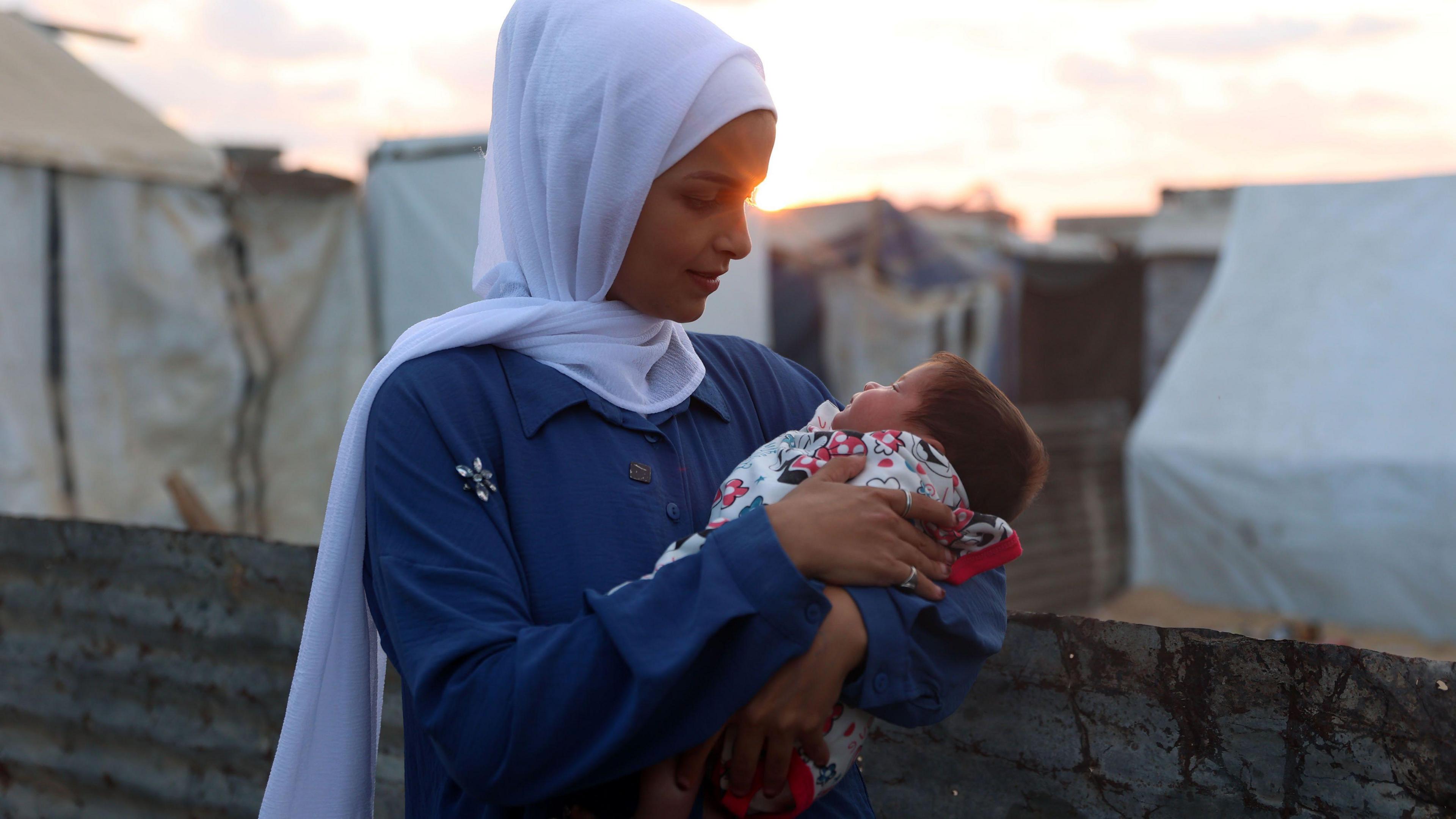 Rana, who is wearing a white hijab and blue dress, looks at her daughter's face while holding the baby in her arms. They are standing in front of tents while the sun is setting behind them.