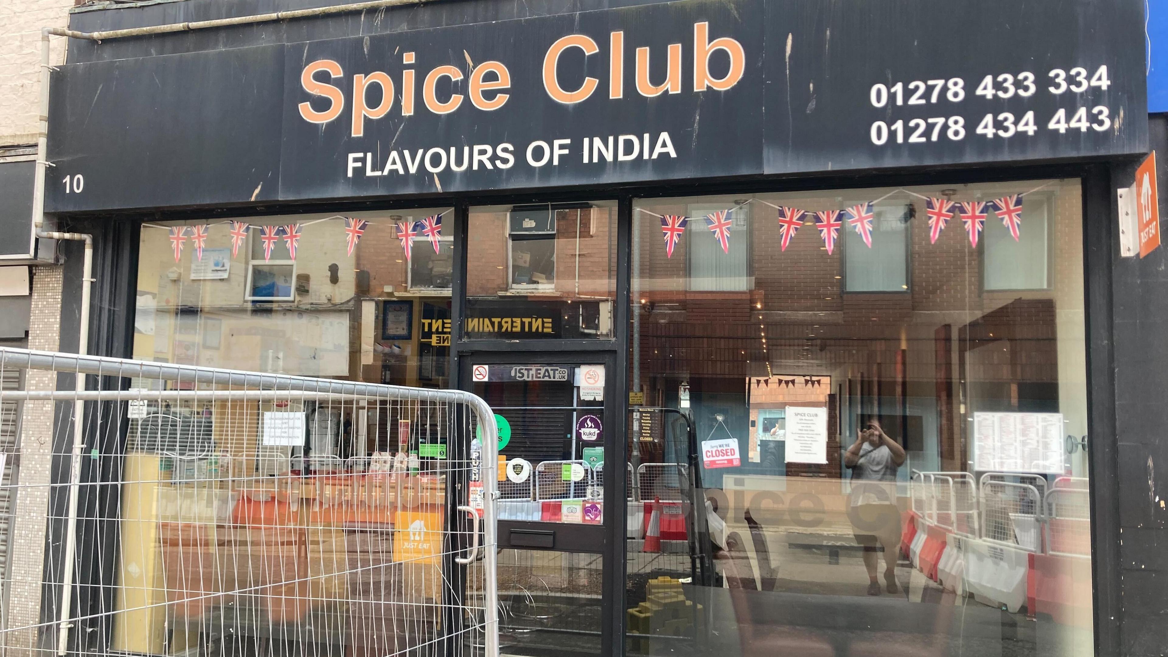 The picture shows the outside of The Spice Club in Bridgwater. The sign is black with Spice Club written in orange paint. The sign also says Flavours of India.