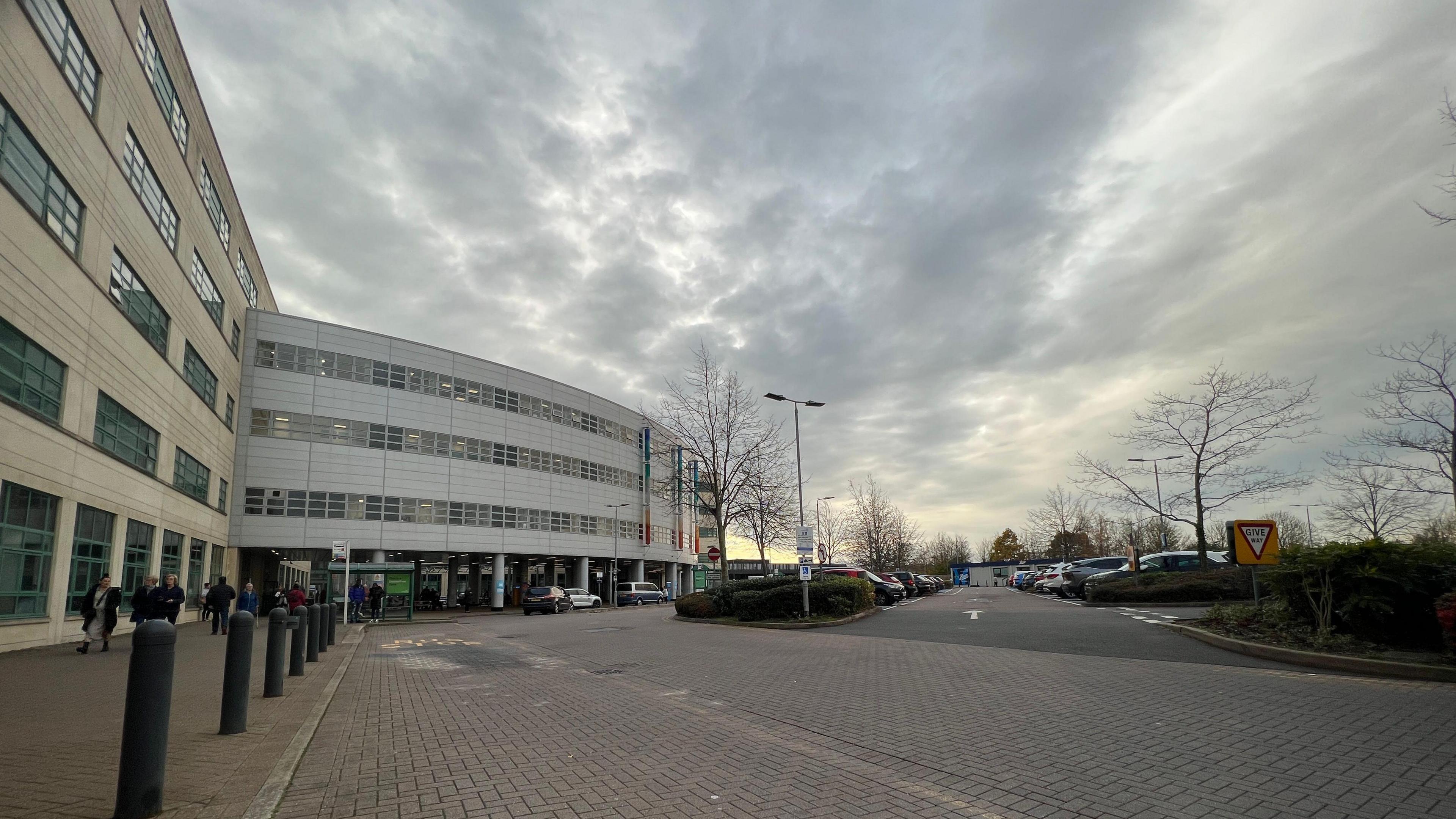 A view of a hospital building with a car park to the right side. It is a cloudy day.