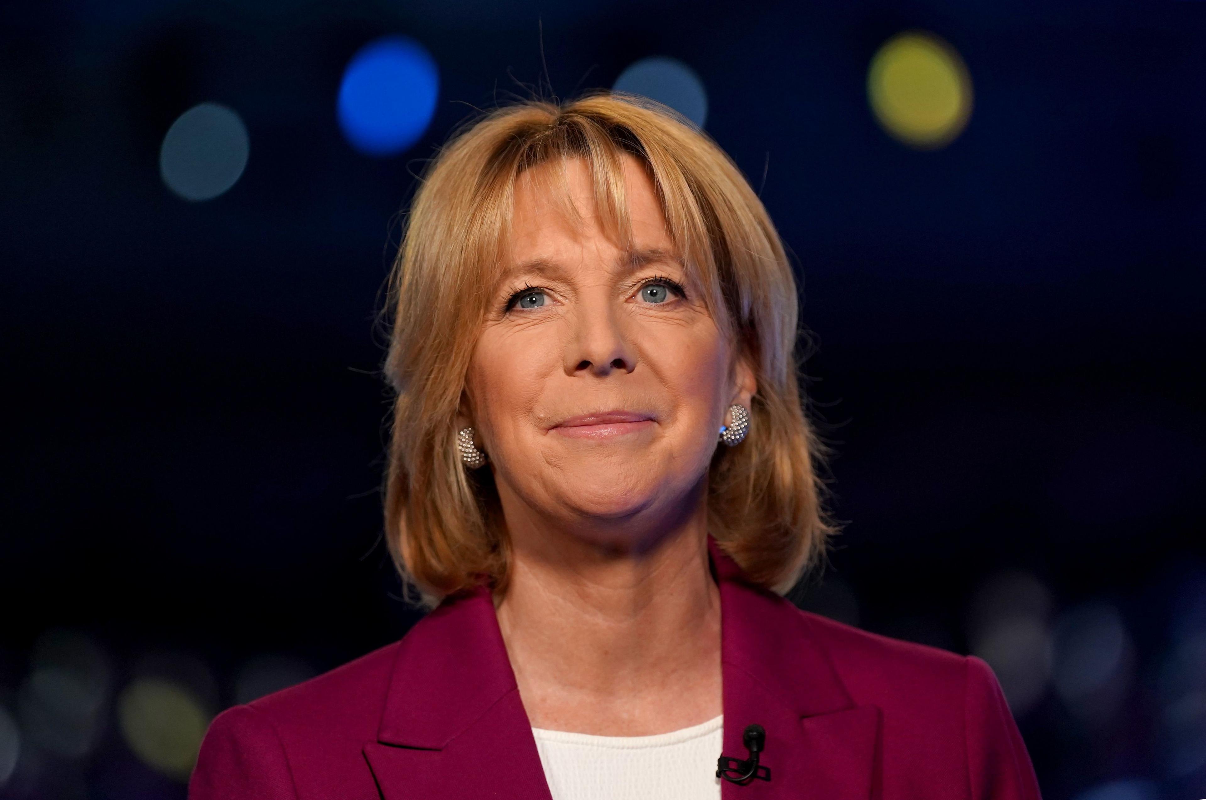 Hazel Irvine smiling gently and looking up with a blurred tv studio background. She is wearing a white t-shirt, sparkly earrings and a deep pink tailored jacket.