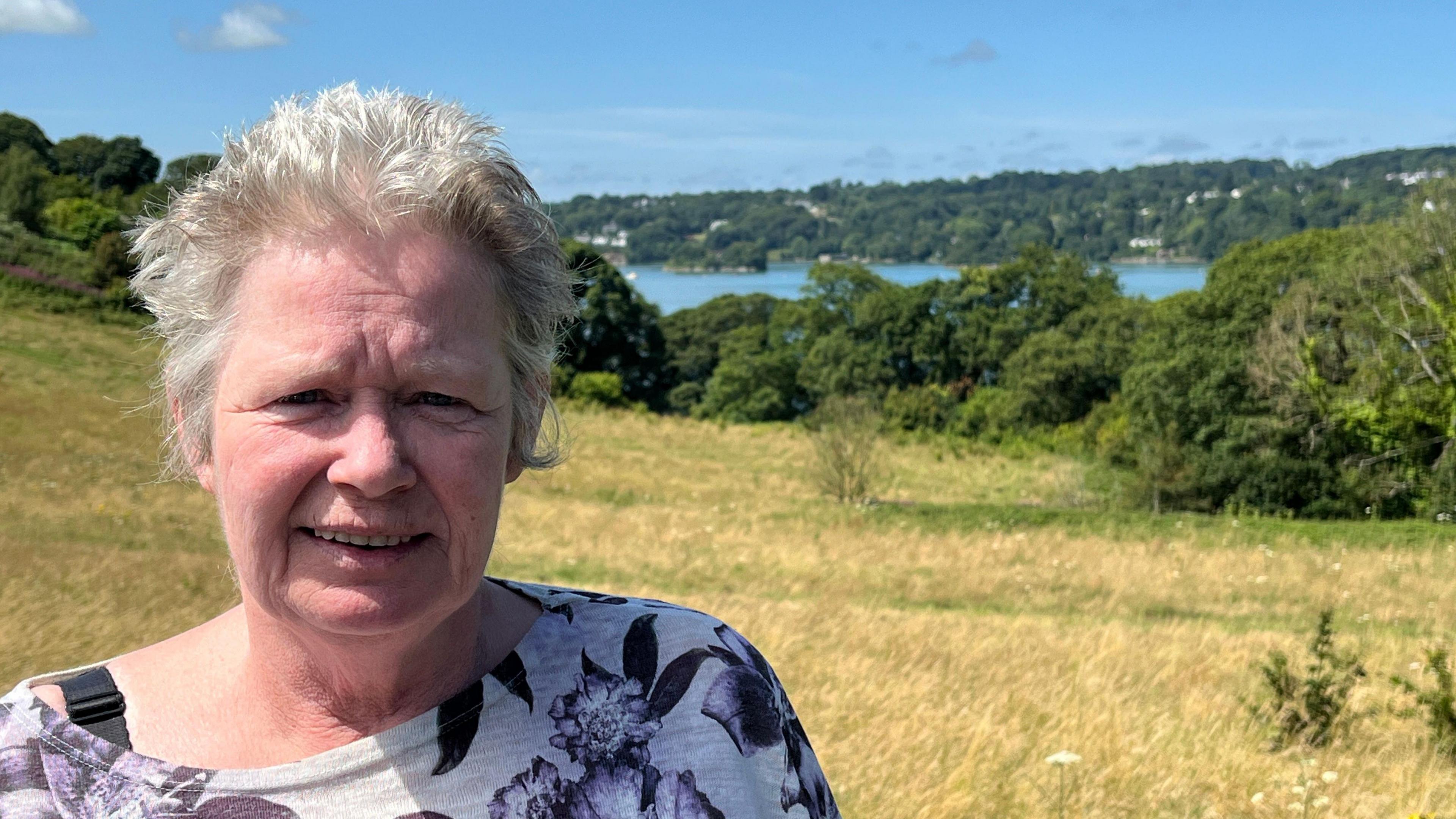 Jan Underwood looking at the camera, she is wearing a purple and white top and behind her is the sea and fields