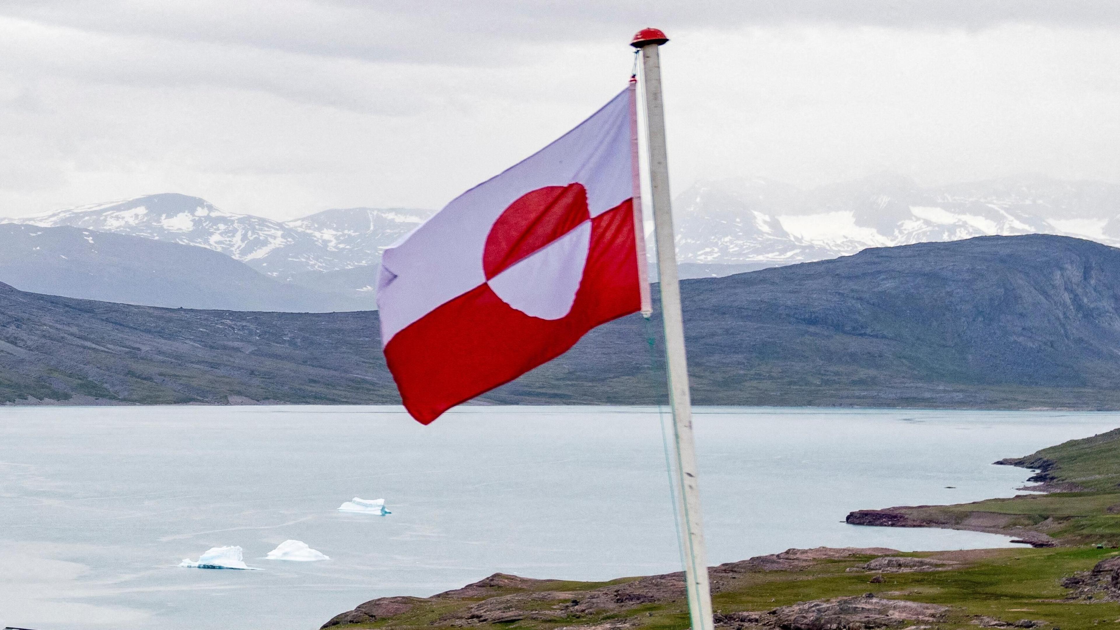 Greenland's flag flies against a backdrop of water and snow-covered mountains. The flag's top half is white and the bottom half is red, with a horizontal line running through it and bisecting a red-and-white circle.