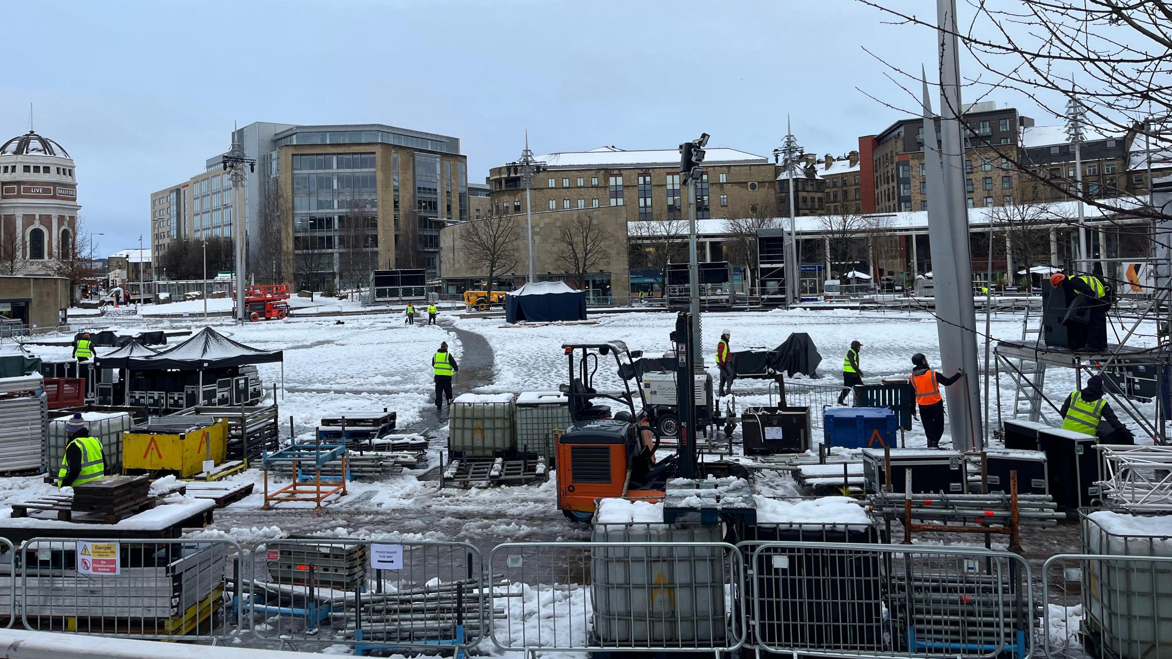 A city square, covered in snow, with various machines, workers and equipment in view. The surrounding cityscape is in the background. Workers wear high vis clothing.