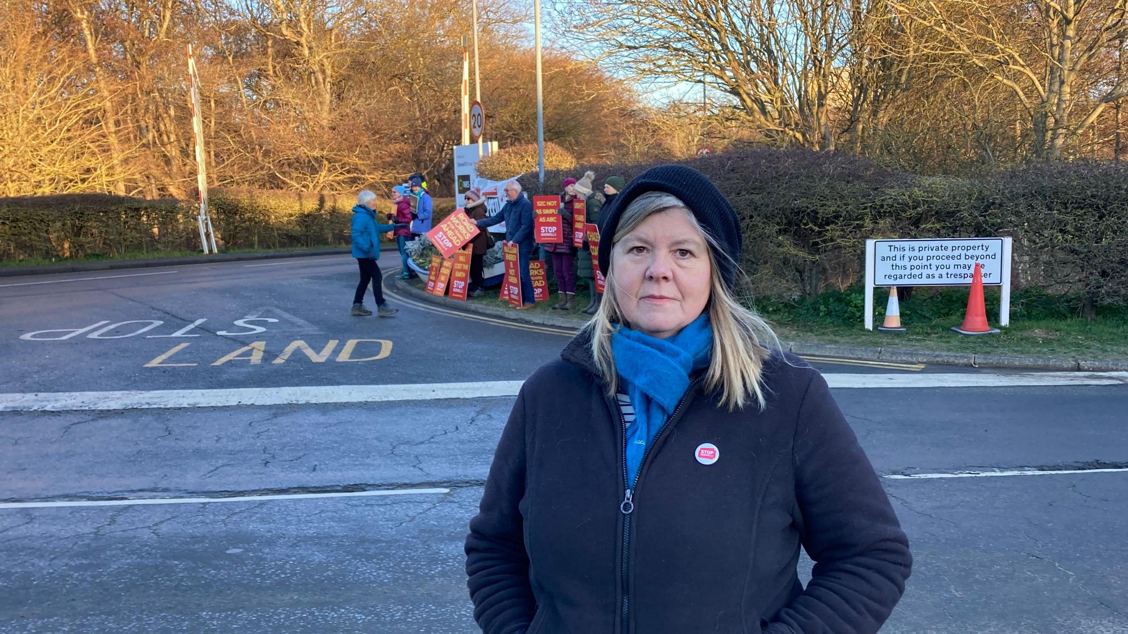 Alison Downes standing outside the Sizewell site. She is in a dark coloured fleece, blue scarf and a dark coloured hat. Behind her are protestors holding  placards citing 'stop sizewell C'. 