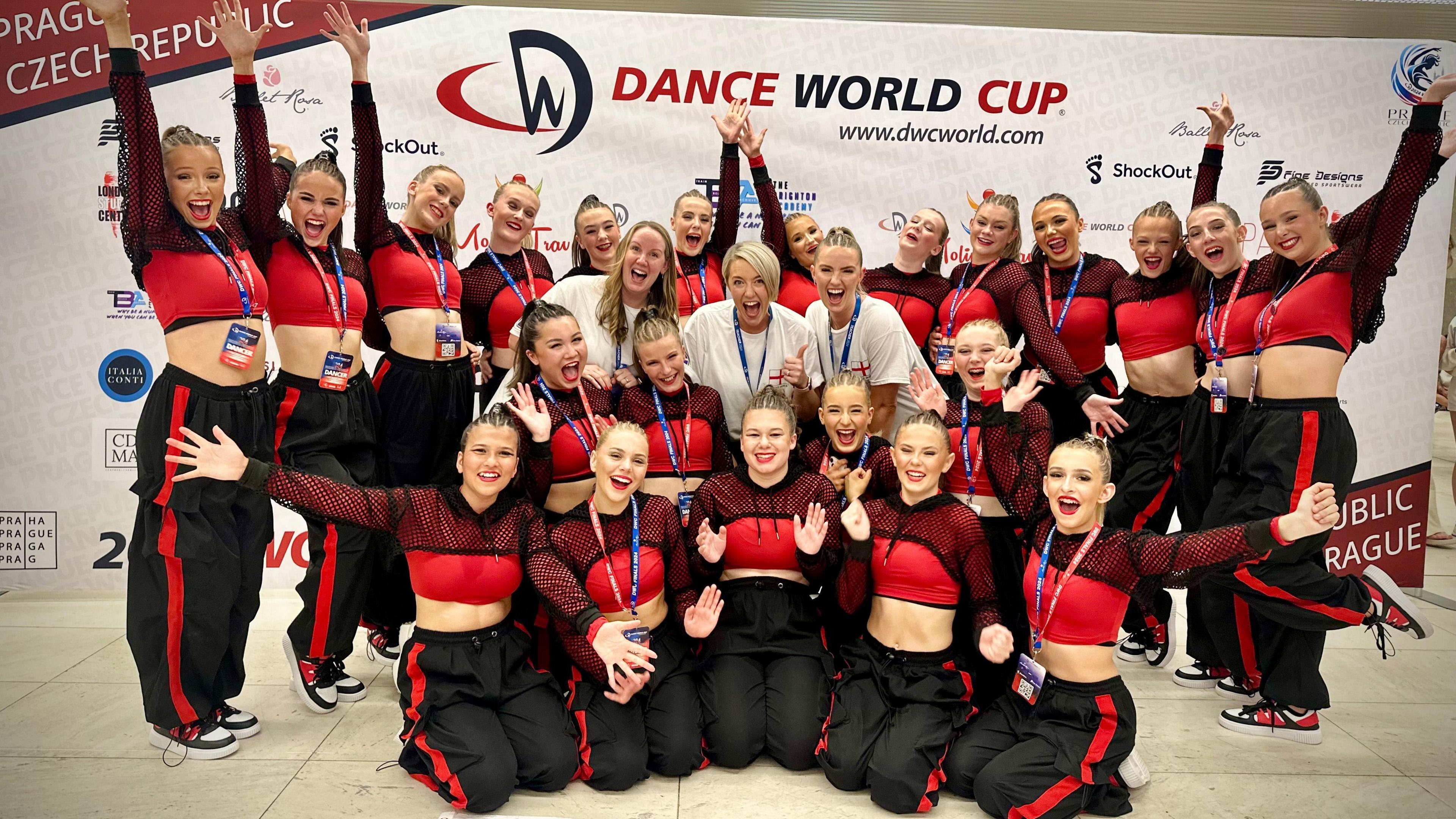 22 dancers pictured all wearing red cropped tops with black trousers a red stripe down the leg, three women in white t-shirts are in the centre. Most of the dancers have their arms up in the air. 
