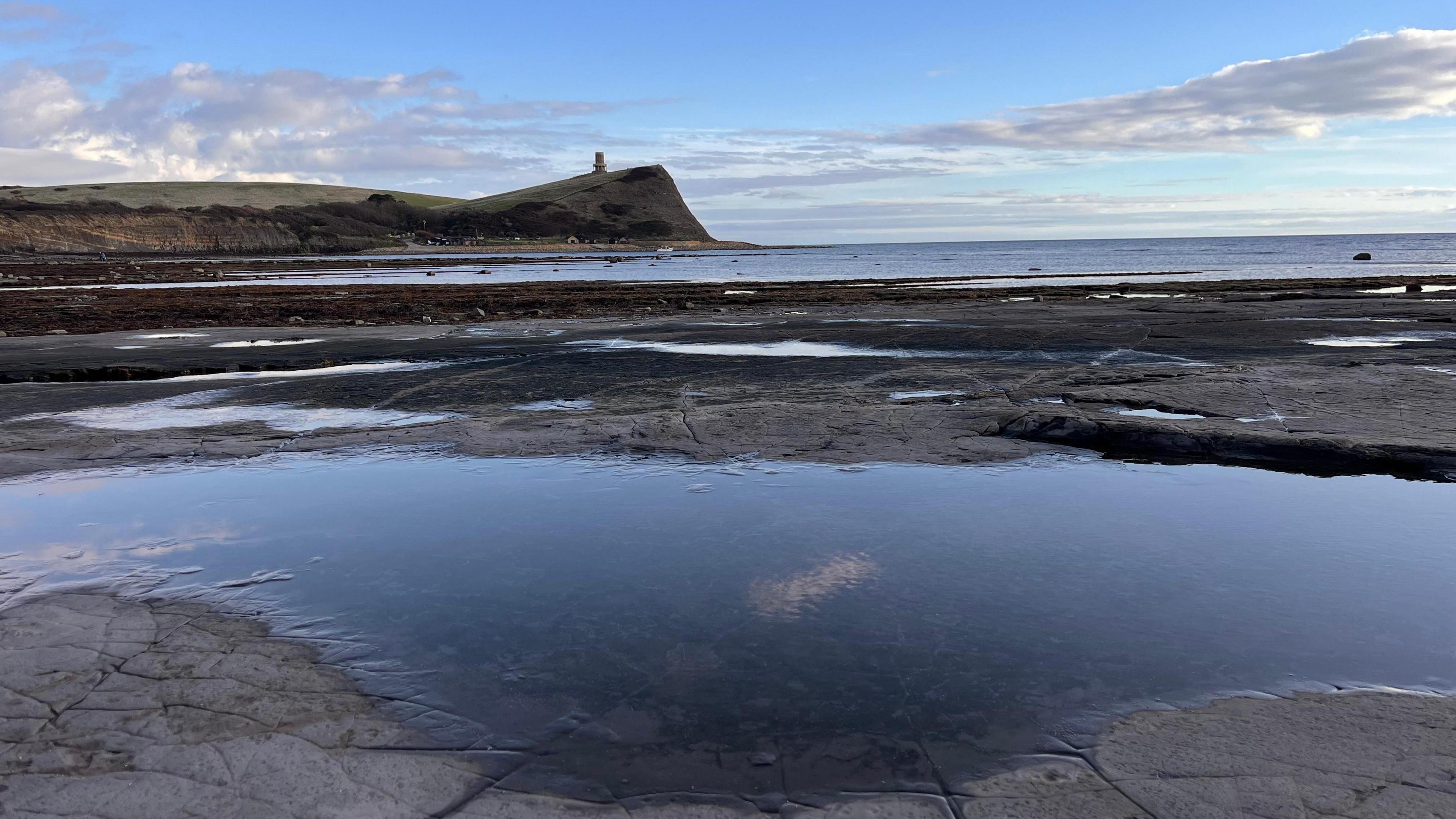 A low-tide reveals a rock pool with the sea visible in the background and the arching coastline emerging from the left