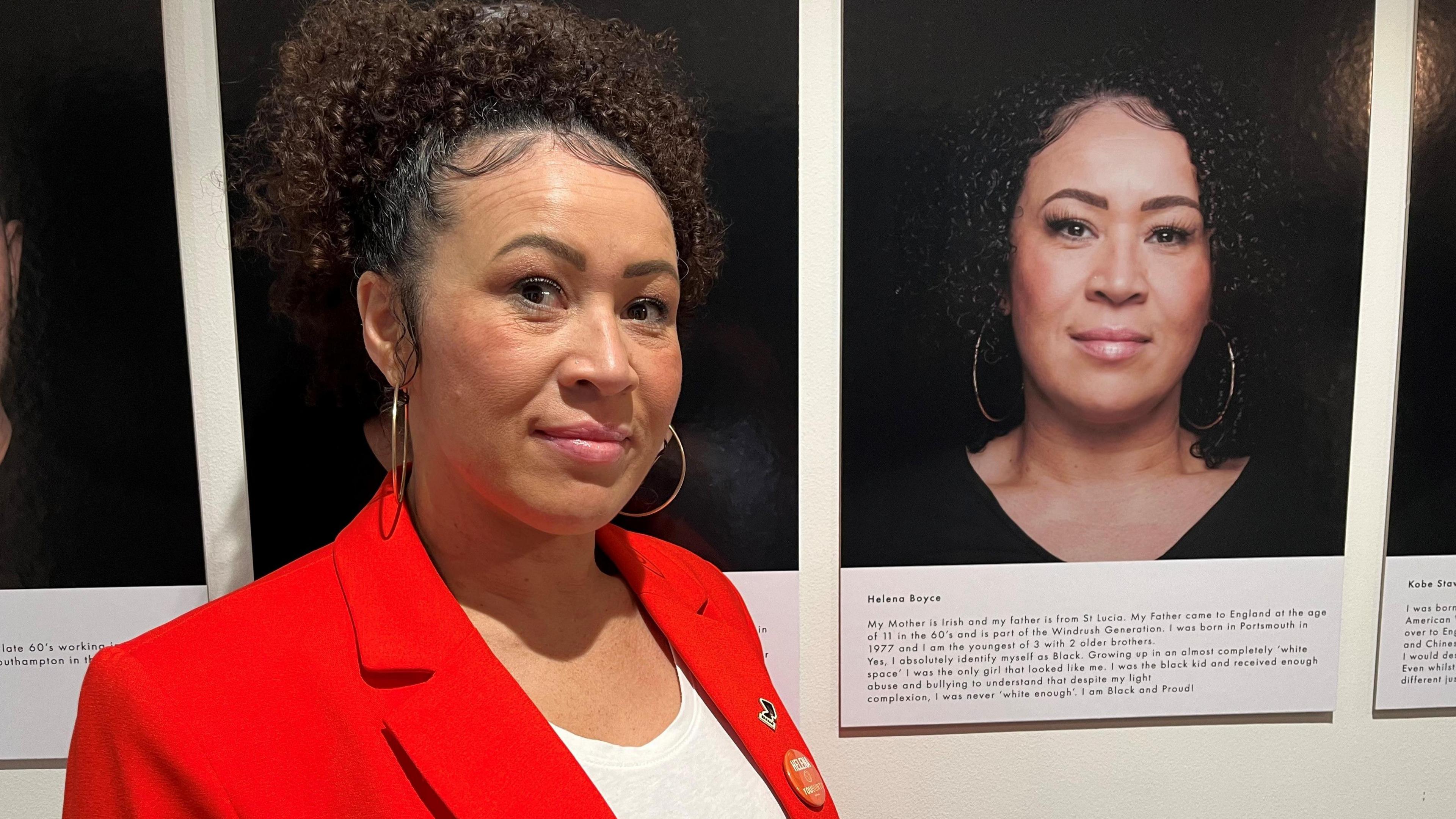 Helena stands to the left of a portrait image of herself. She is wearing a red blazer and white t-shirt and has her hair piled on top of her head and wears large slim gold hoop earrings. She is looking into the camera with a small smile on her face.