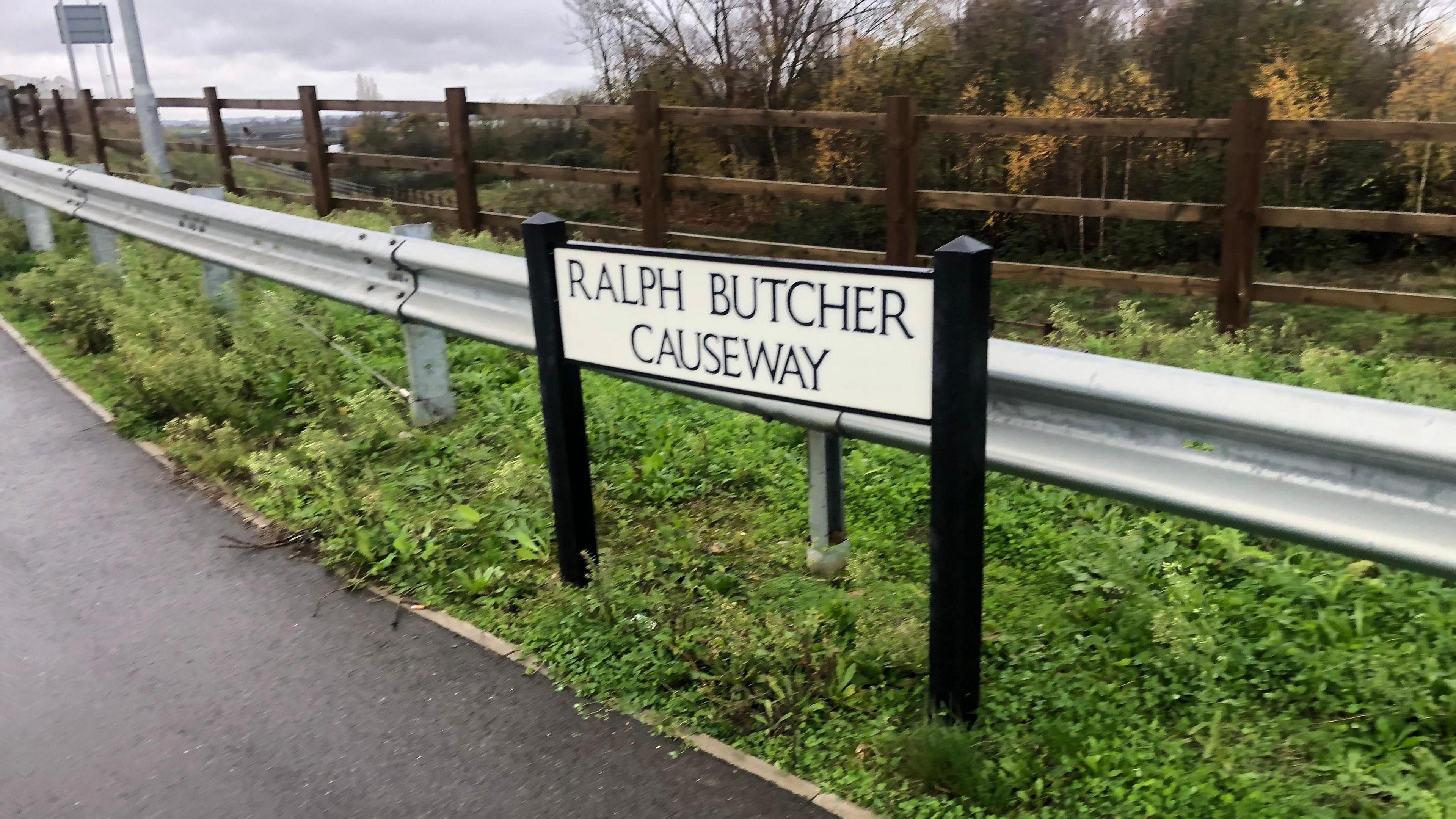 The side of a bridge with a silver railing and a black and white sign that reads "Ralph Butcher Causeway"