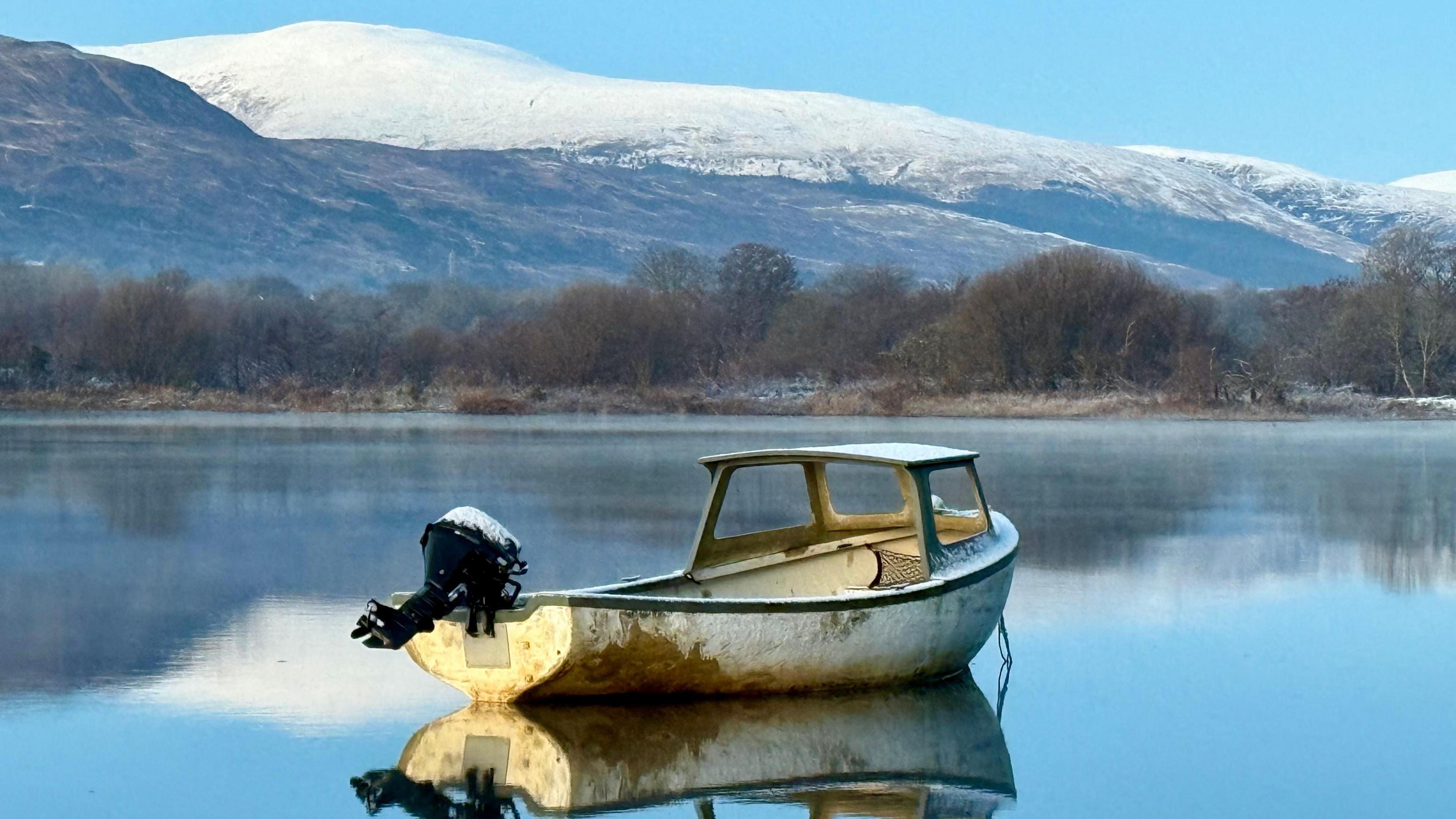 A small white boat sits in a loch in front of some snowy mountains. The water is so calm the boat and mountains are reflected onto the water.