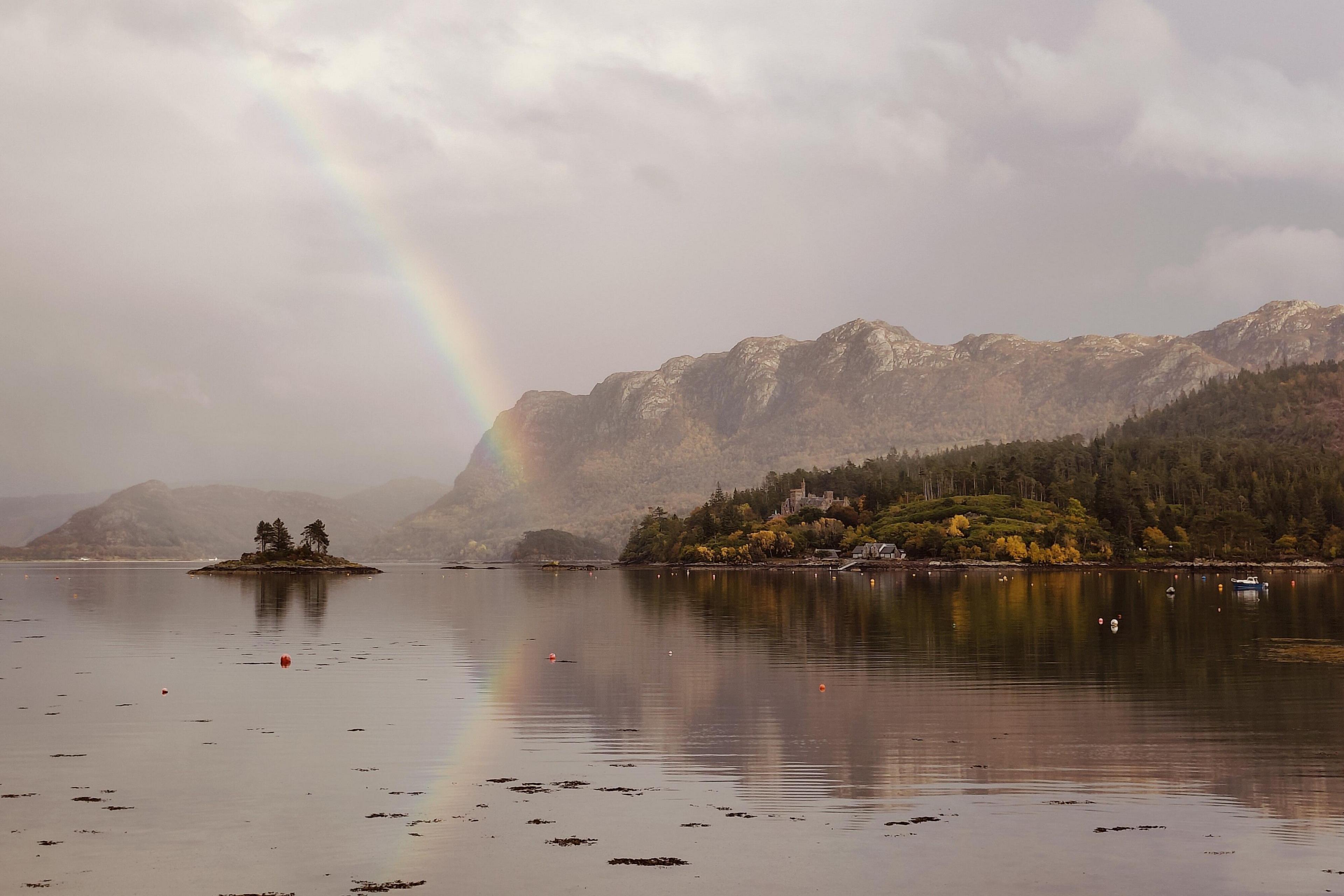 An atmospheric and hazy looking Highland scene with mountains in the background and water in the foreground. There is also a rinbow.