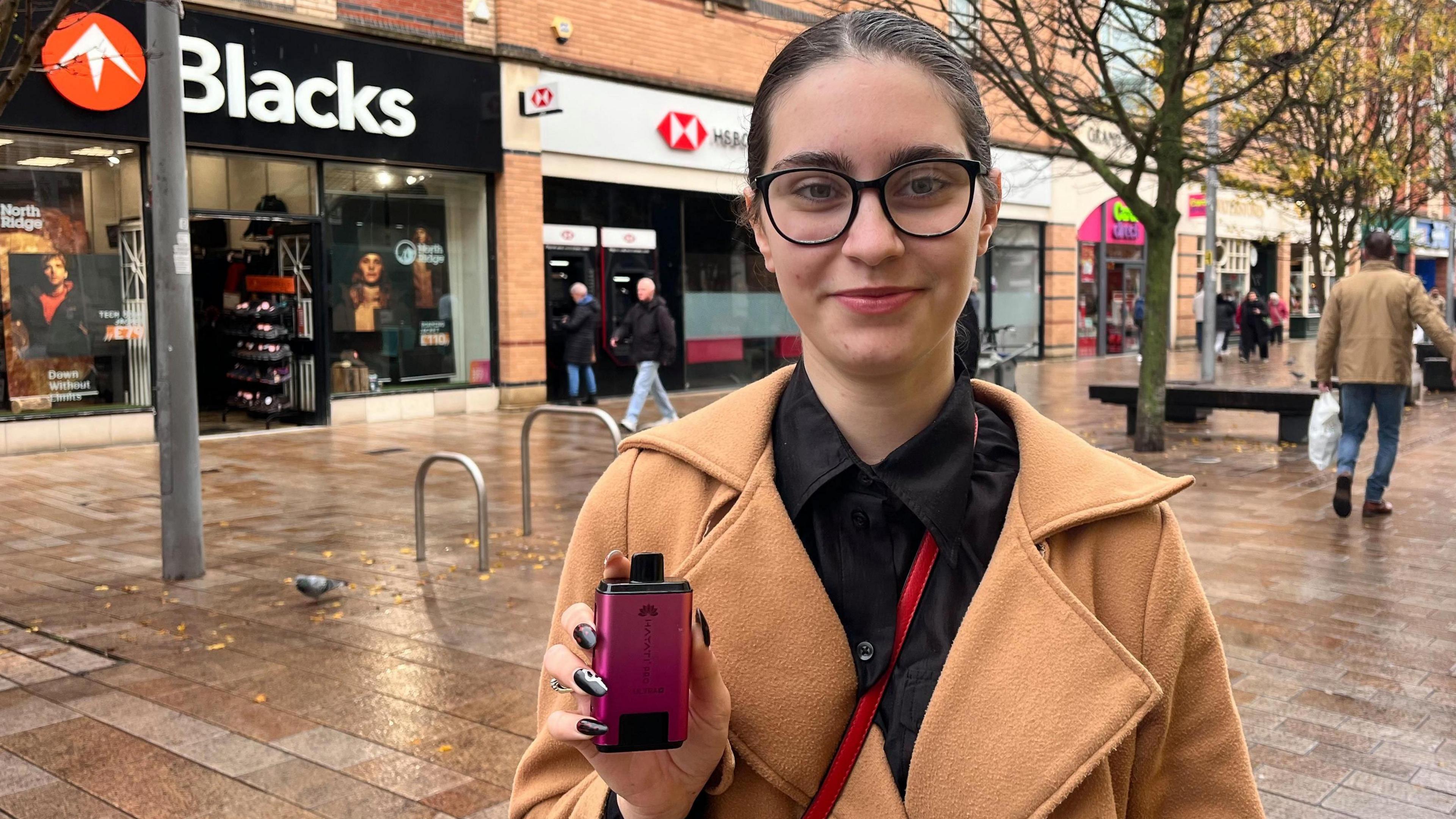 A 20-year-old woman with black hair and wearing black-rimmed glasses, a camel coat with wide lapels and a black shirt holds up a pink vape as she stands in a pedestrian street in front of a row of shops.
