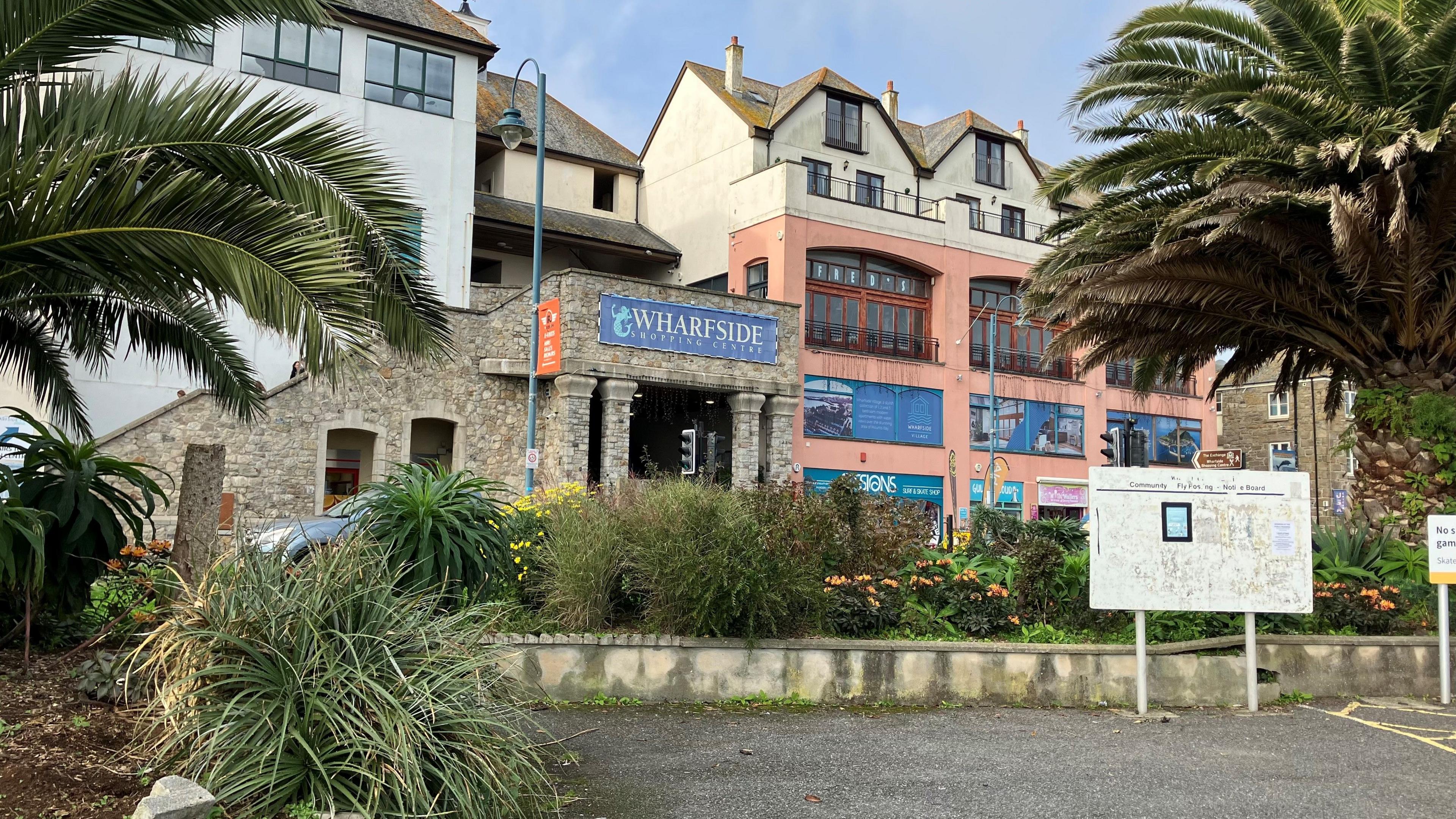 The façade of the Wharfside building is made of granite and it is nestled within other buildings. A car park and palm tree are in the foreground.