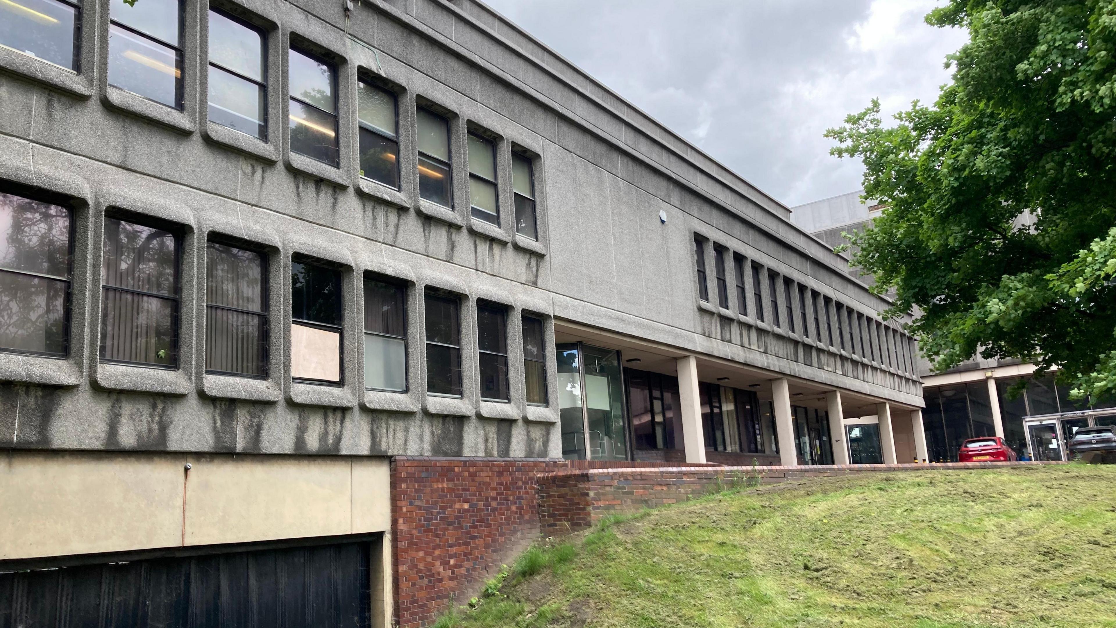 A grey 1960s brutalist building, situated next to a grassy patch of land. 