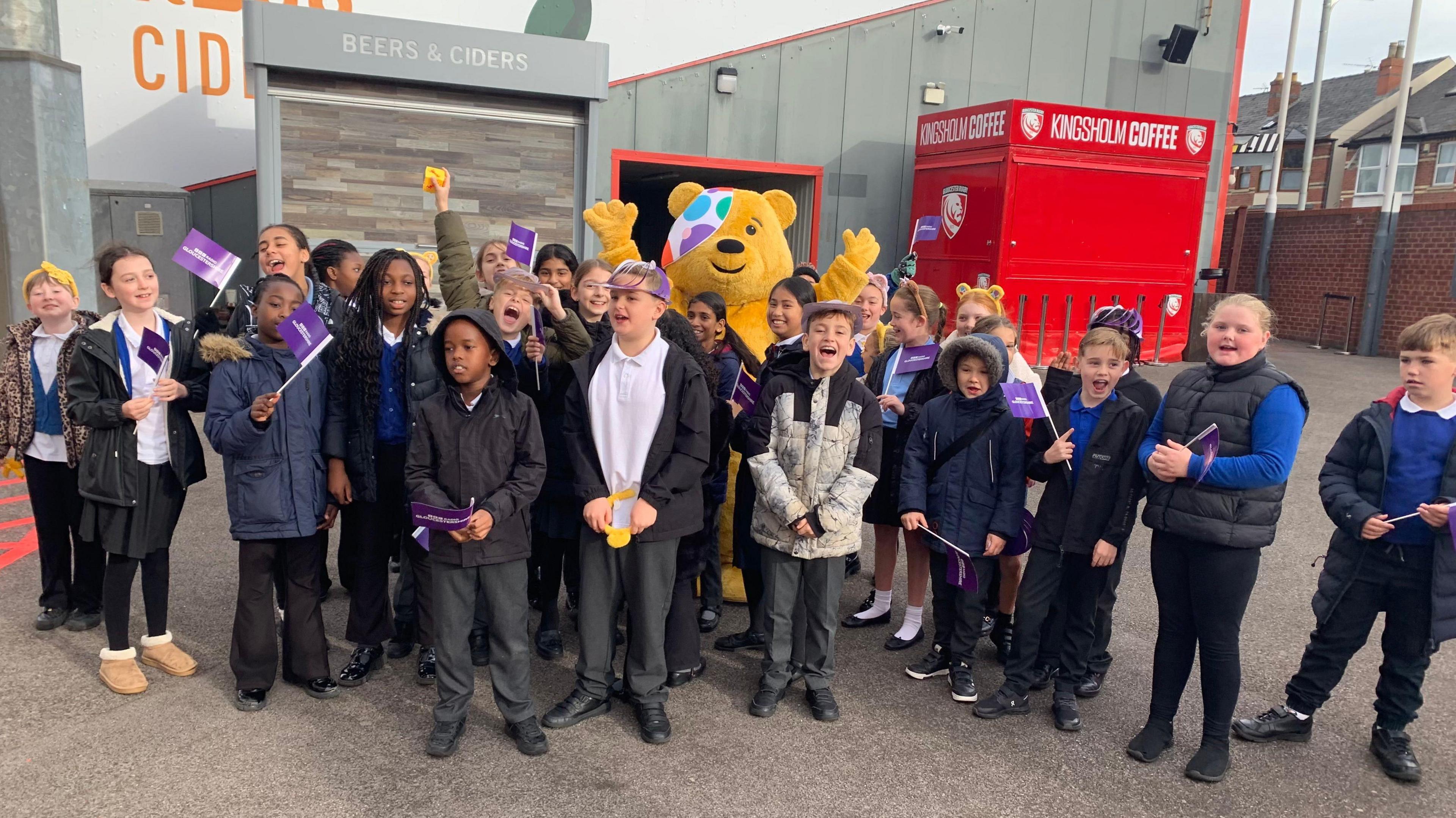 A group of school children are gathered at Kingsholm stadium. They are waving purple branded BBC Gloucestershire flags and Pudsey the bear is in the background of the huddle.